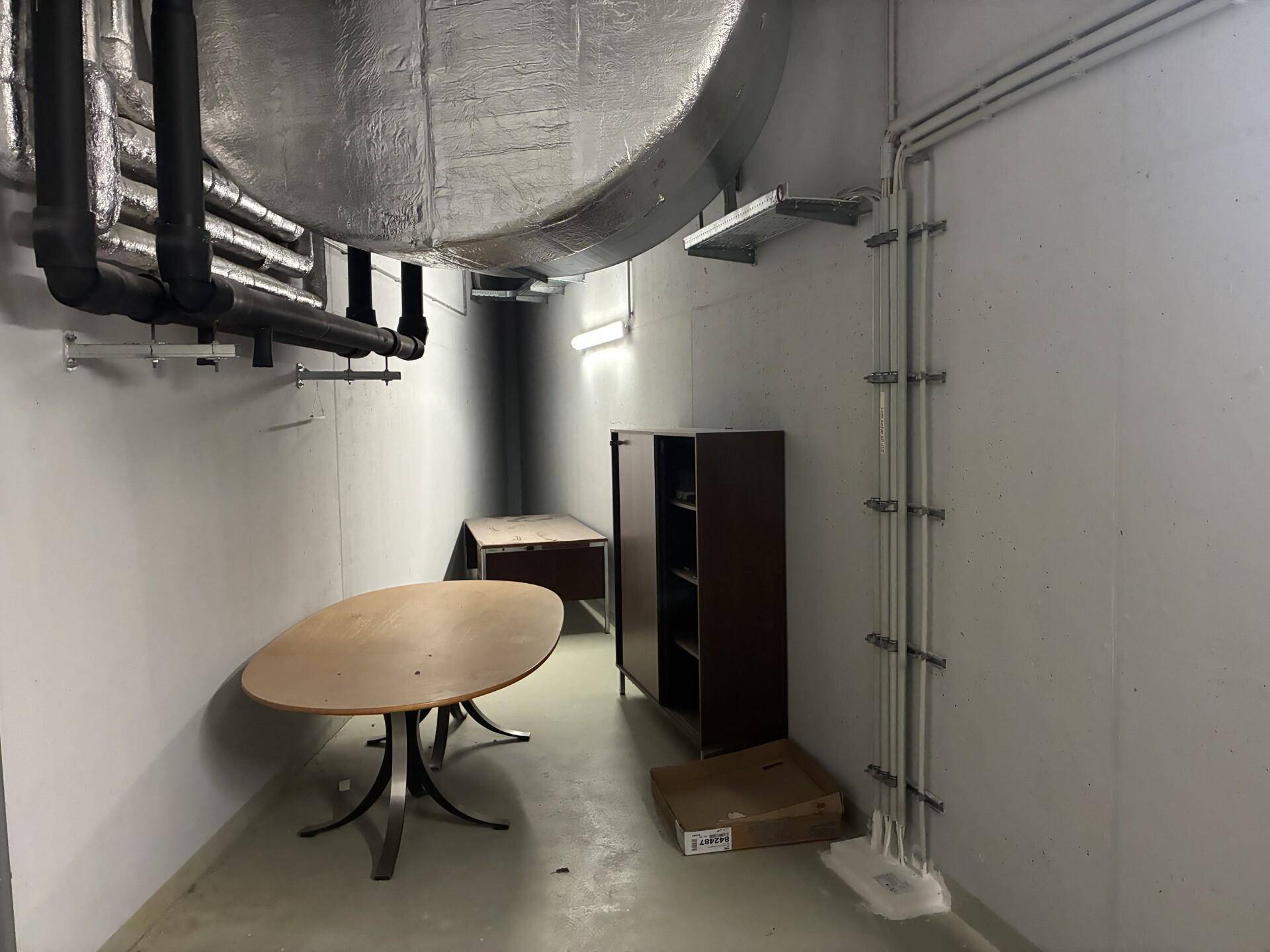 Technikraum mit sichtbaren Rohren, Lüftungskanälen und einem Betonsockel, funktional eingerichtet.