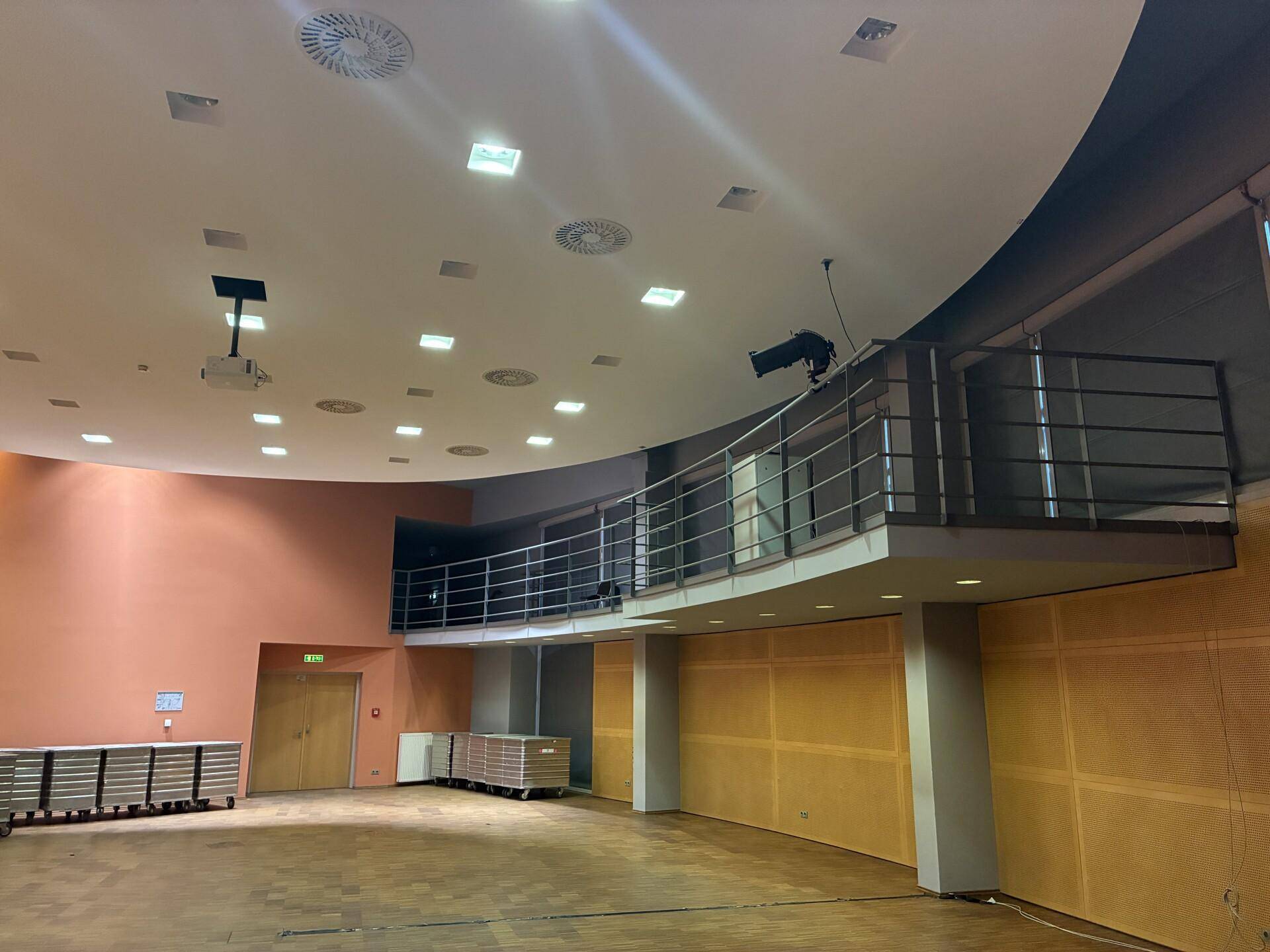 Weitläufiger Konferenzsaal mit einer geschwungenen Empore und Holzboden, bereit für Veranstaltungen.