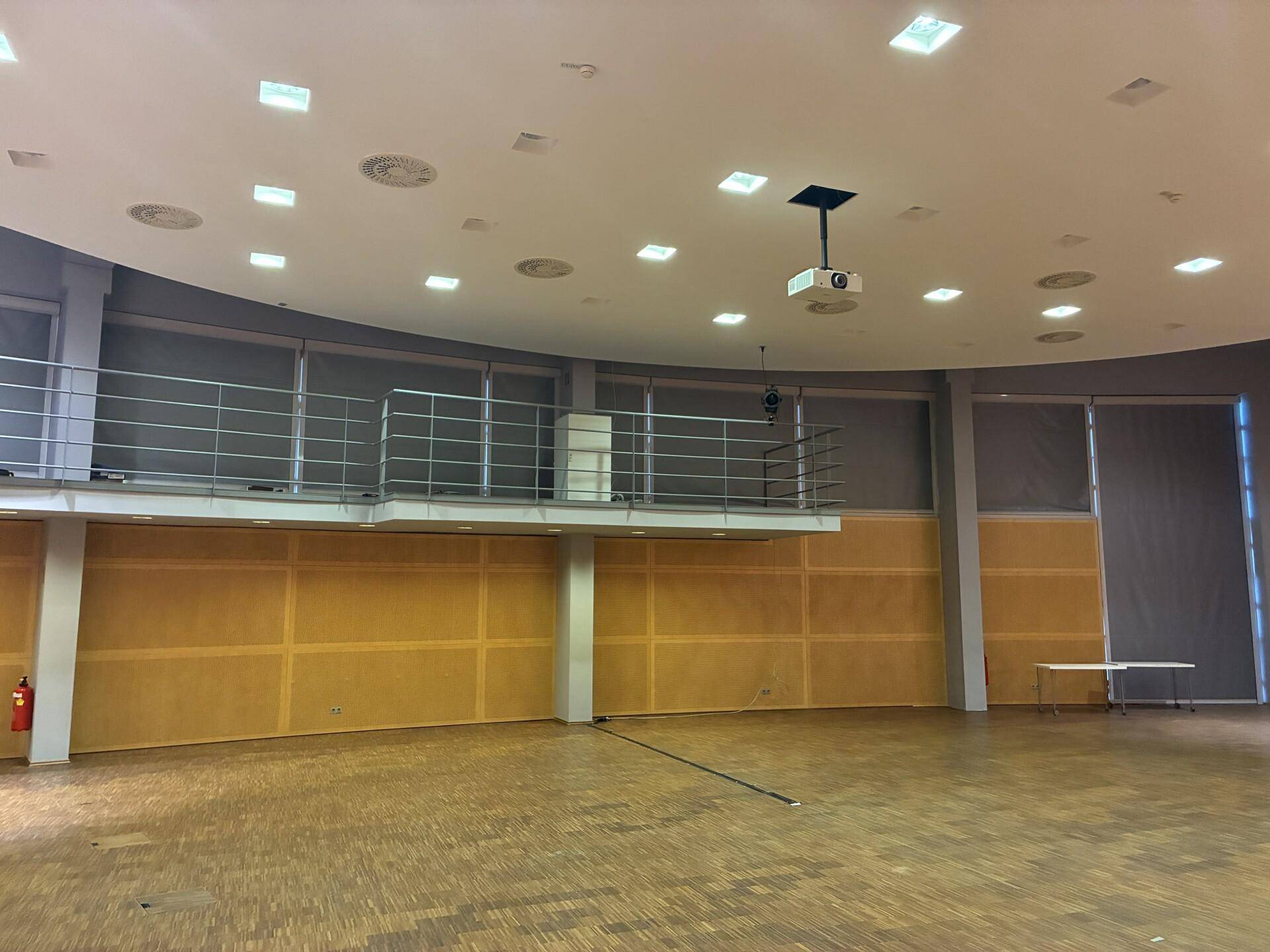 Großer Veranstaltungsraum mit einer Empore, Holzparkettboden und Projektor an der Decke für Events.