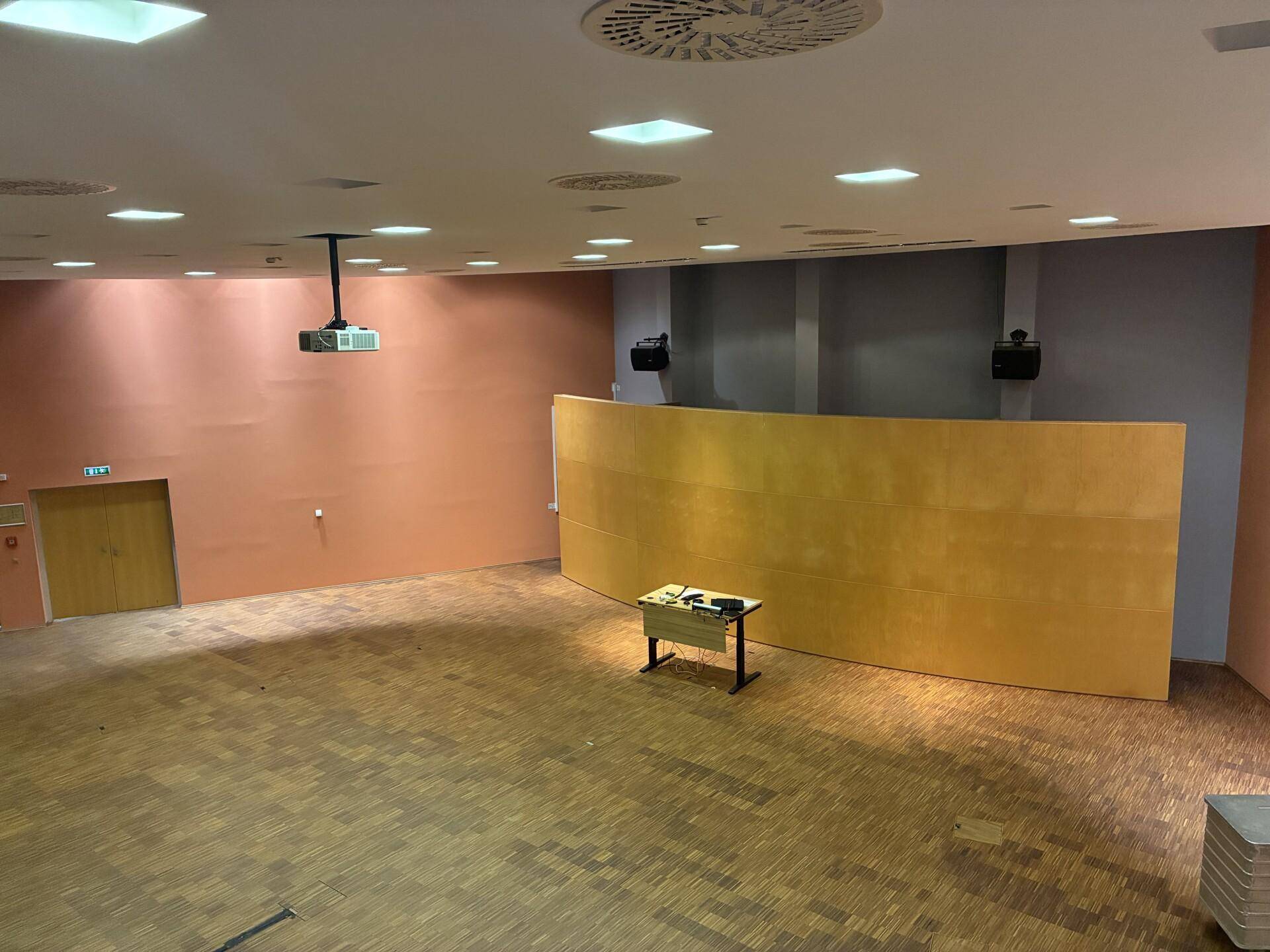Großer Konferenzraum mit Holzparkettboden, Projektor und einer geschwungenen Holzwand für Präsentationen.