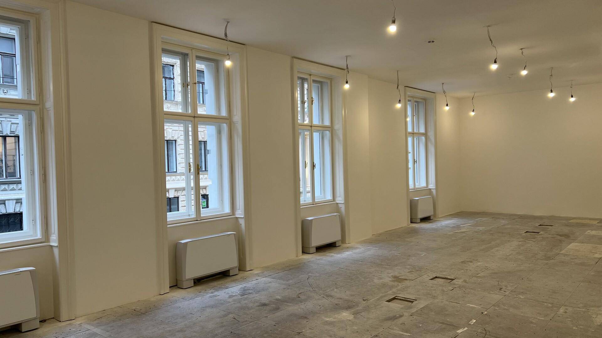 Heller Büroraum mit mehreren hohen Fenstern, die viel Tageslicht hereinlassen, und einem Betonboden.
