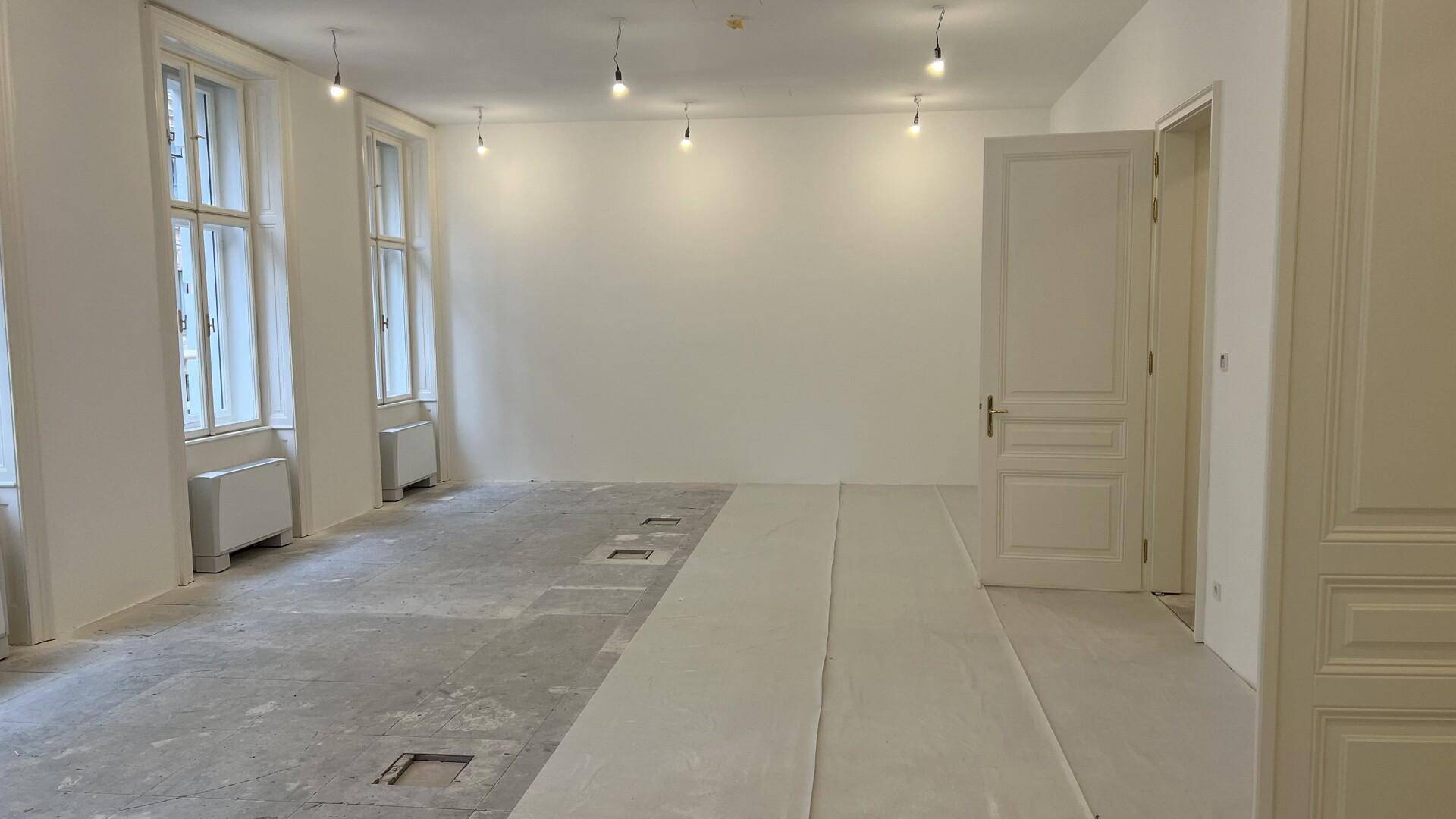 Geräumiger, unmöblierter Raum mit hohen Fenstern und freiliegendem Betonboden, bereit für den Ausbau.