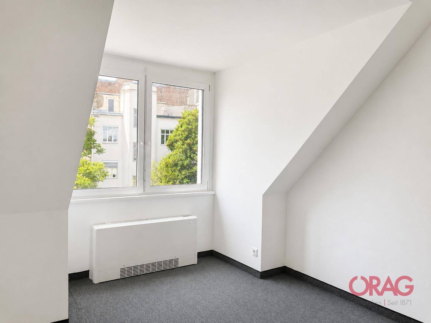 Kleiner Büroraum im Dachgeschoss mit schräger Decke, Teppichboden und einem Fenster mit Blick ins Grüne.