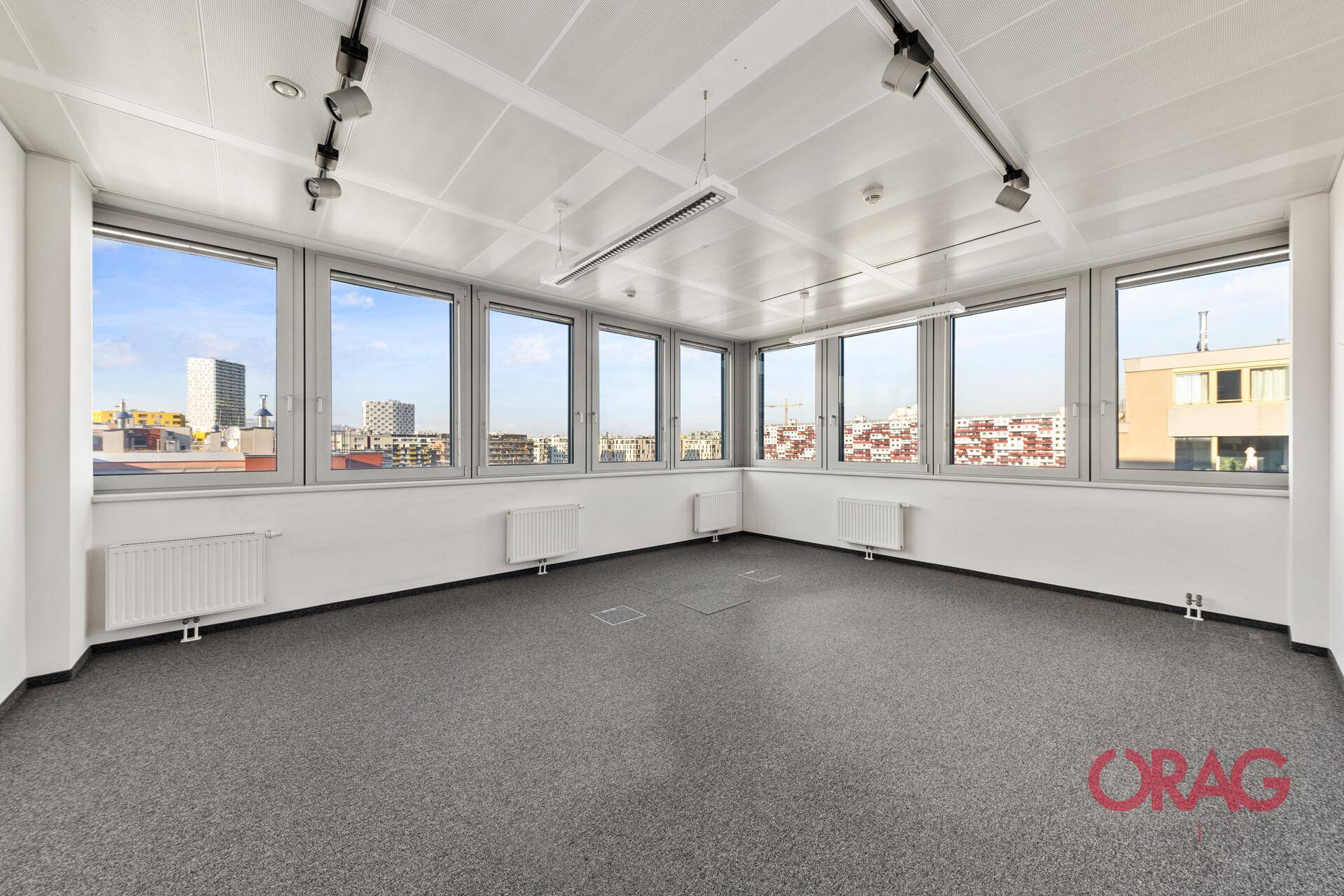 Leeres Büro mit Teppichboden, großen Fenstern und Blick auf die Stadtlandschaft.