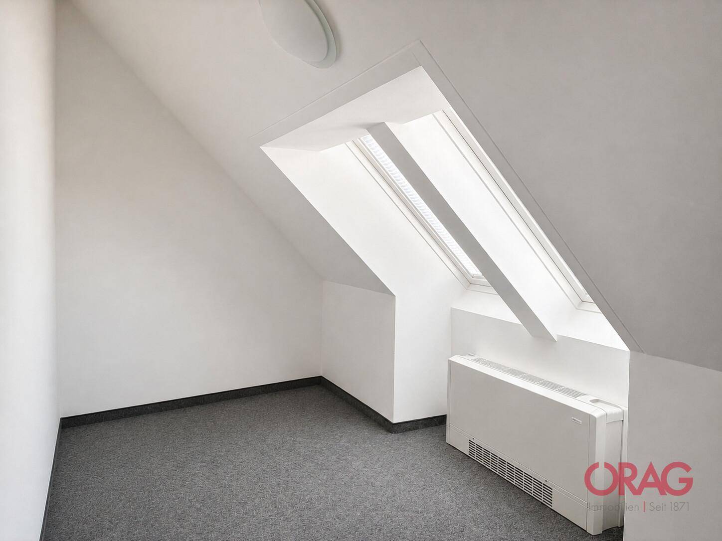 Heller Dachbodenraum mit schrägen Wänden, einem Dachfenster und grauem Teppichboden, ideal als Büro.