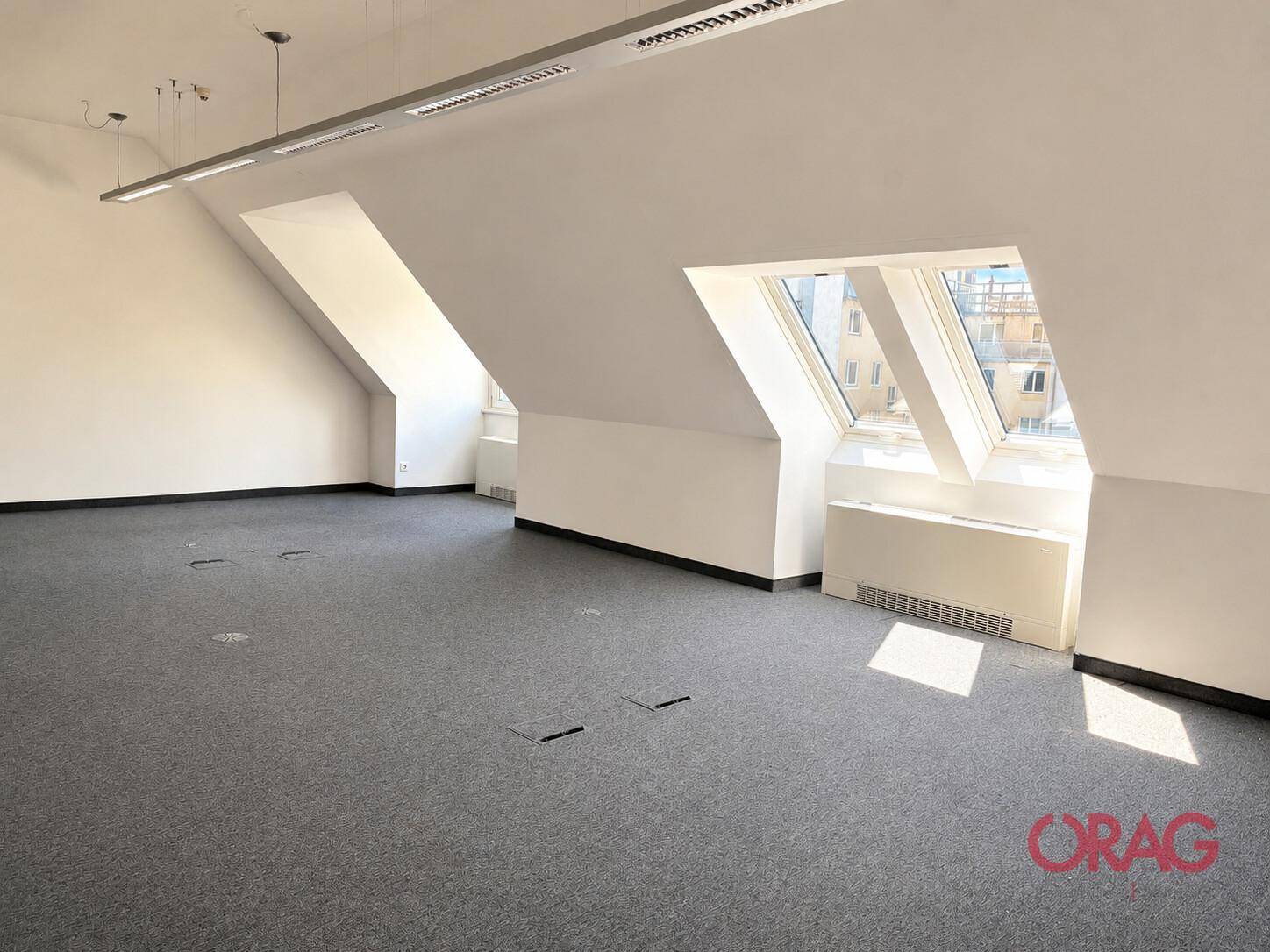Großer, heller Büroraum im Dachgeschoss mit schrägen Decken, grauem Teppichboden und integrierten Bodensteckdosen.