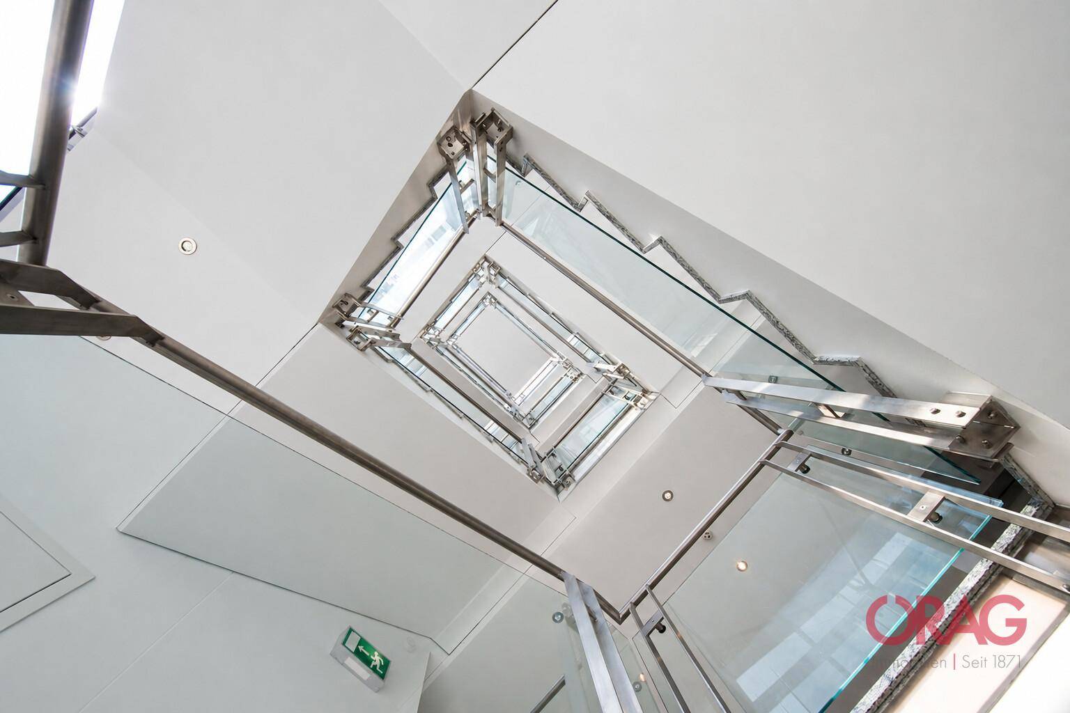 Blick nach oben in ein modernes Treppenhaus mit Glasgeländern und Edelstahlakzenten, das viel Licht hereinlässt.