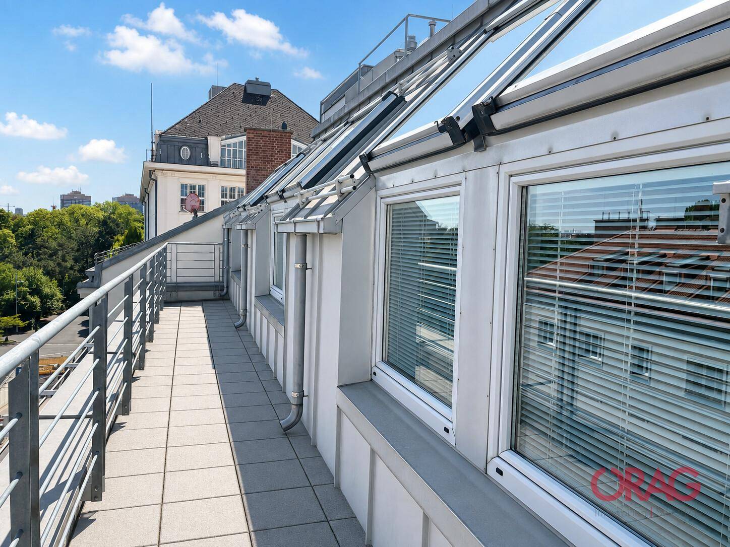 Lange, schmale Terrasse im Obergeschoss mit Fliesenboden, Metallgeländer und Blick auf die umliegenden Gebäude.