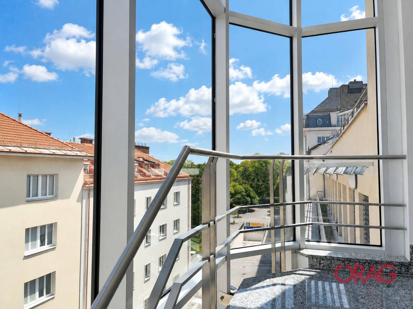 Blick aus einem modernen Treppenhaus mit Glasfassade und Edelstahlgeländer auf die umliegenden Gebäude und die Straße.