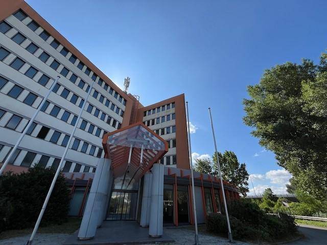 Eingangsbereich eines modernen Bürogebäudes mit Glasvordach und Säulen unter blauem Himmel.