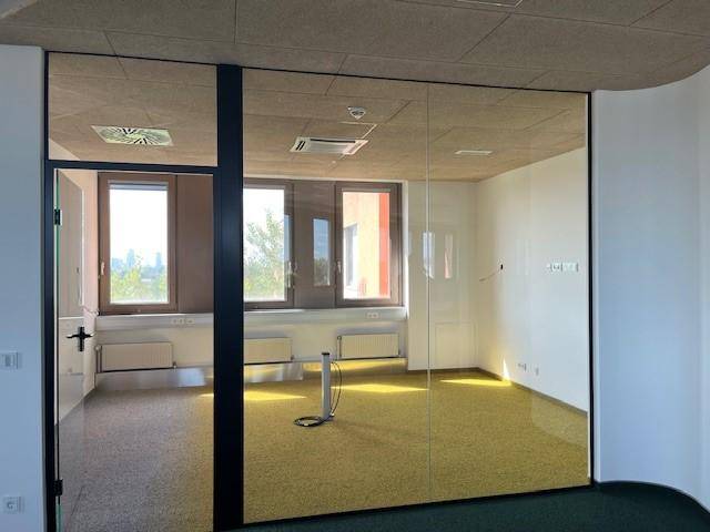 Leeres Büro mit gelbem Teppichboden, großen Fenstern und Heizkörpern, gesehen durch eine Glaswand.