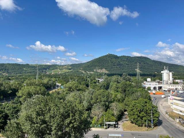 Weitläufiger Blick auf eine grüne Hügellandschaft mit Bäumen, Straßen und einigen Gebäuden unter blauem Himmel.