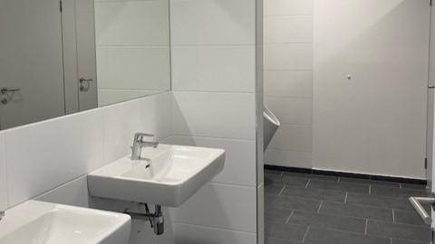 Sauberer öffentlicher Waschraum mit weißen Waschbecken, Spiegeln und einem Urinal, gefliest in Grau und Weiß.