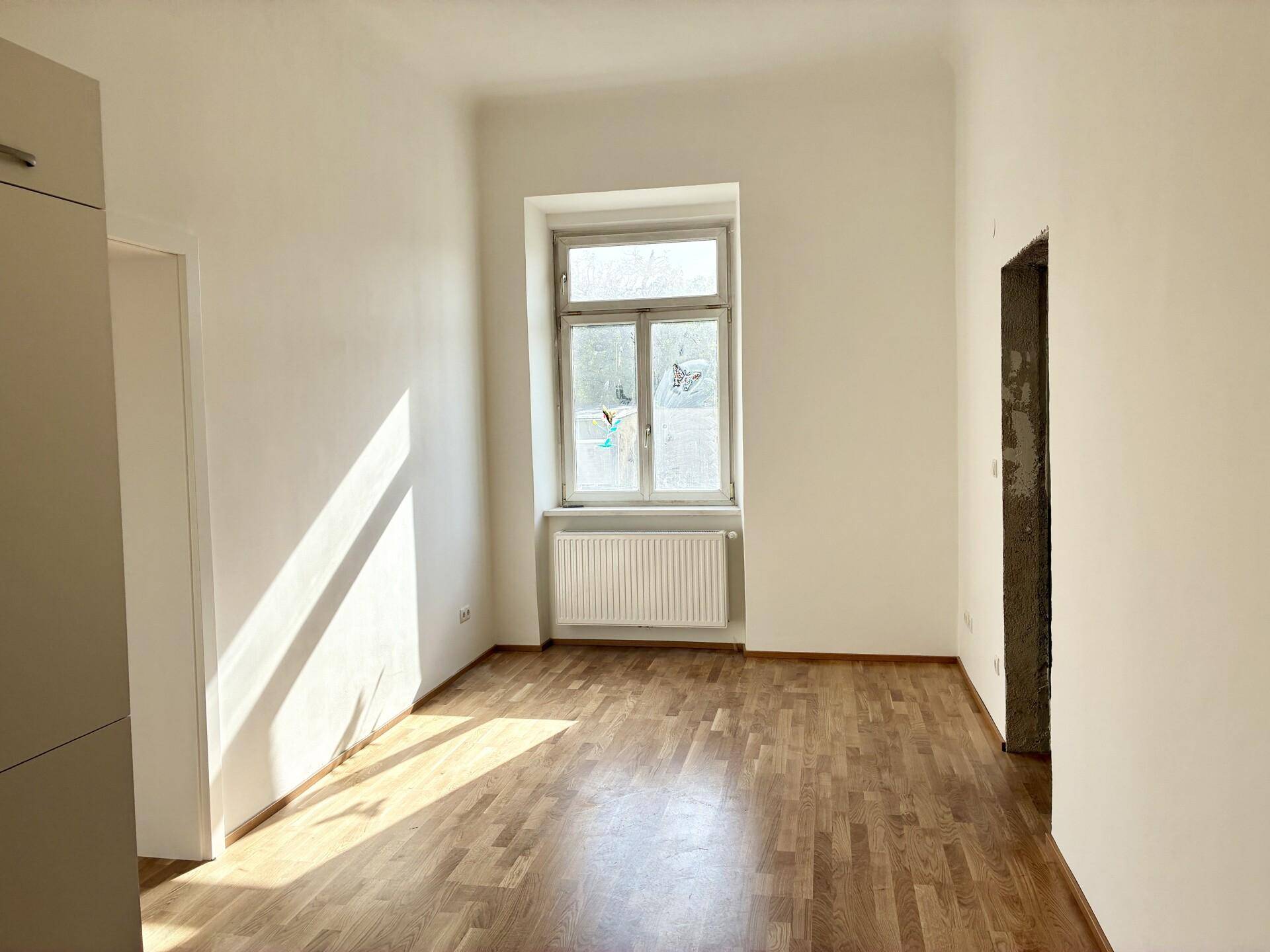 Geräumiges, helles Zimmer mit Holzboden und einem Fenster, das Sonnenlicht in den Raum bringt.