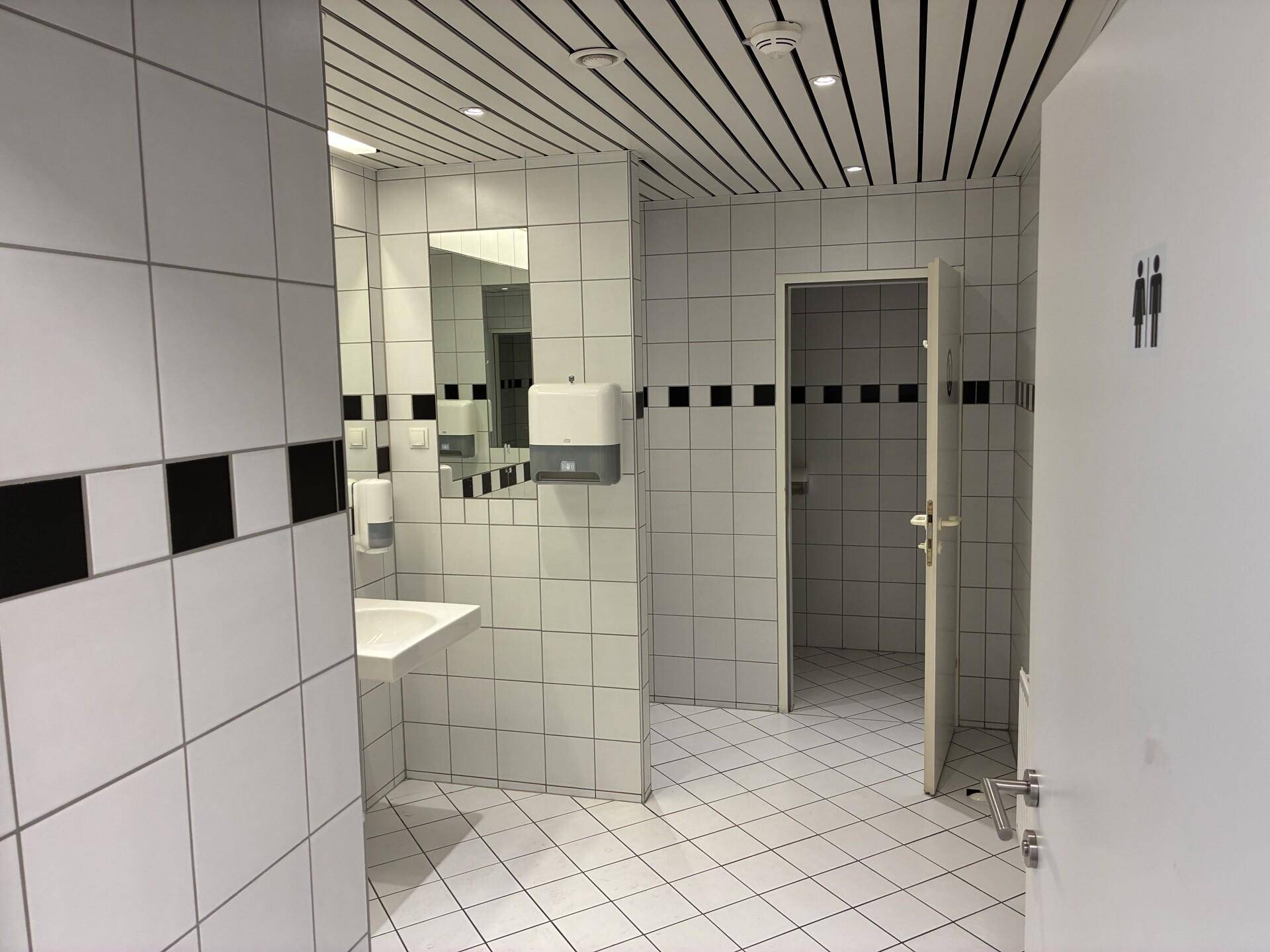 Gefliester Sanitärbereich mit weißen Fliesen, schwarzem Akzentstreifen, Waschbecken und Spiegel.
