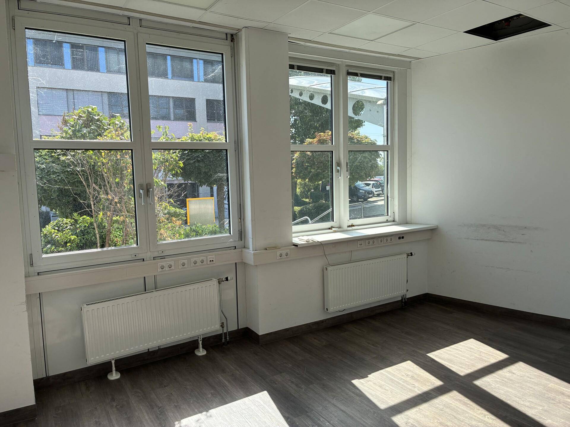 Leeres Büro mit großen Fenstern, die viel Tageslicht hereinlassen, und einem dunklen Laminatboden.