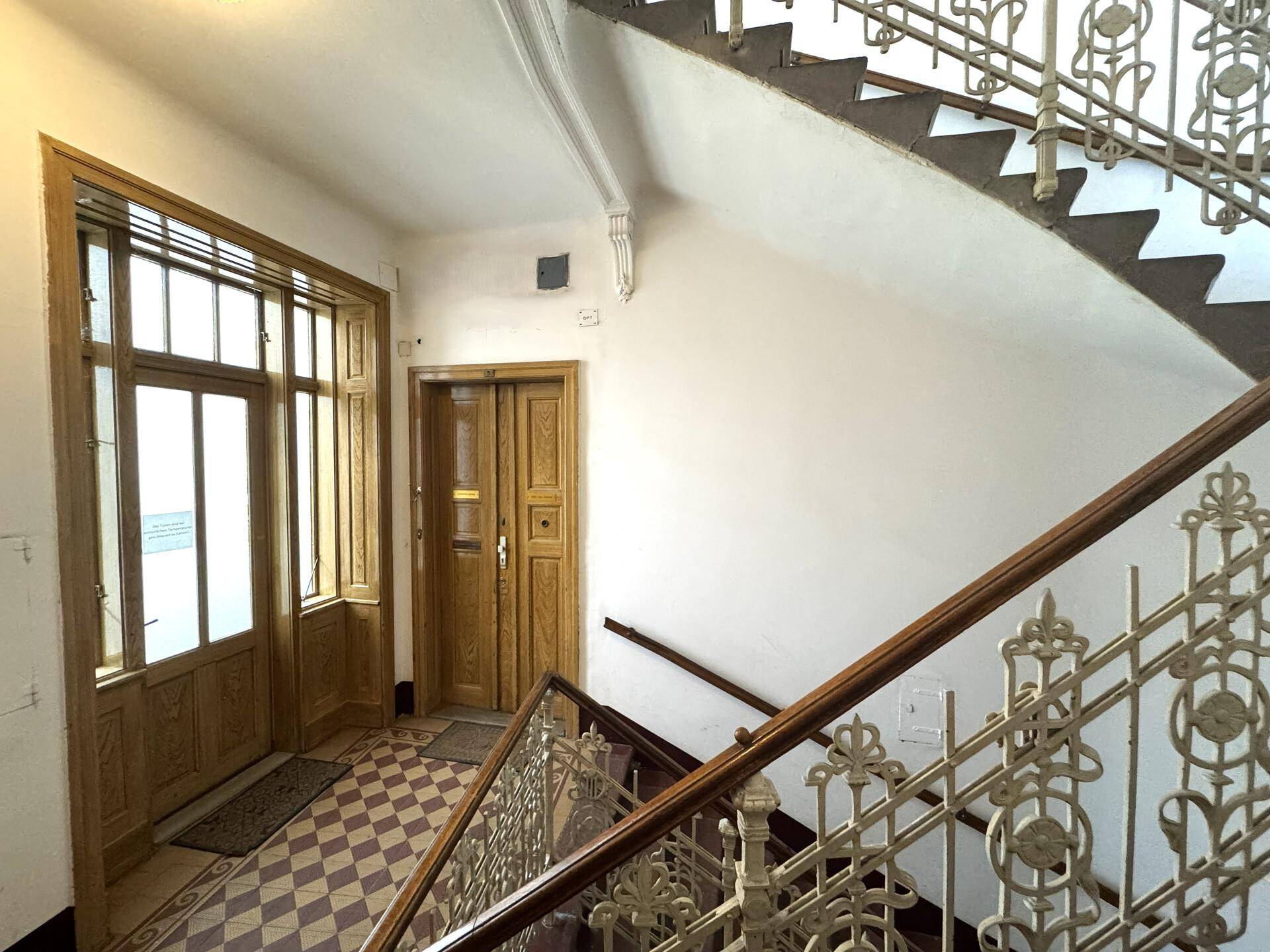 Elegantes Treppenhaus in einem Altbau mit kunstvollem Geländer, Holzstufen und kariertem Fliesenboden im Eingangsbereich.