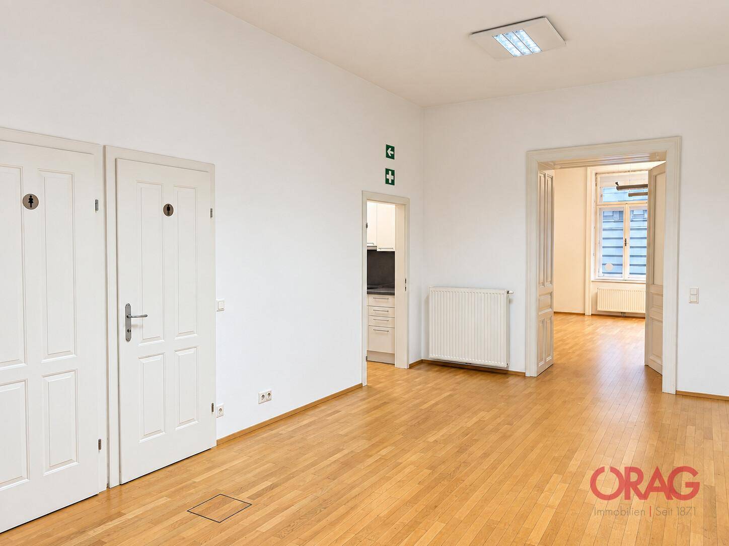 Heller Raum mit weiß gestrichenen Wänden, Holzparkettboden und einem Heizkörper unter einem Fenster.