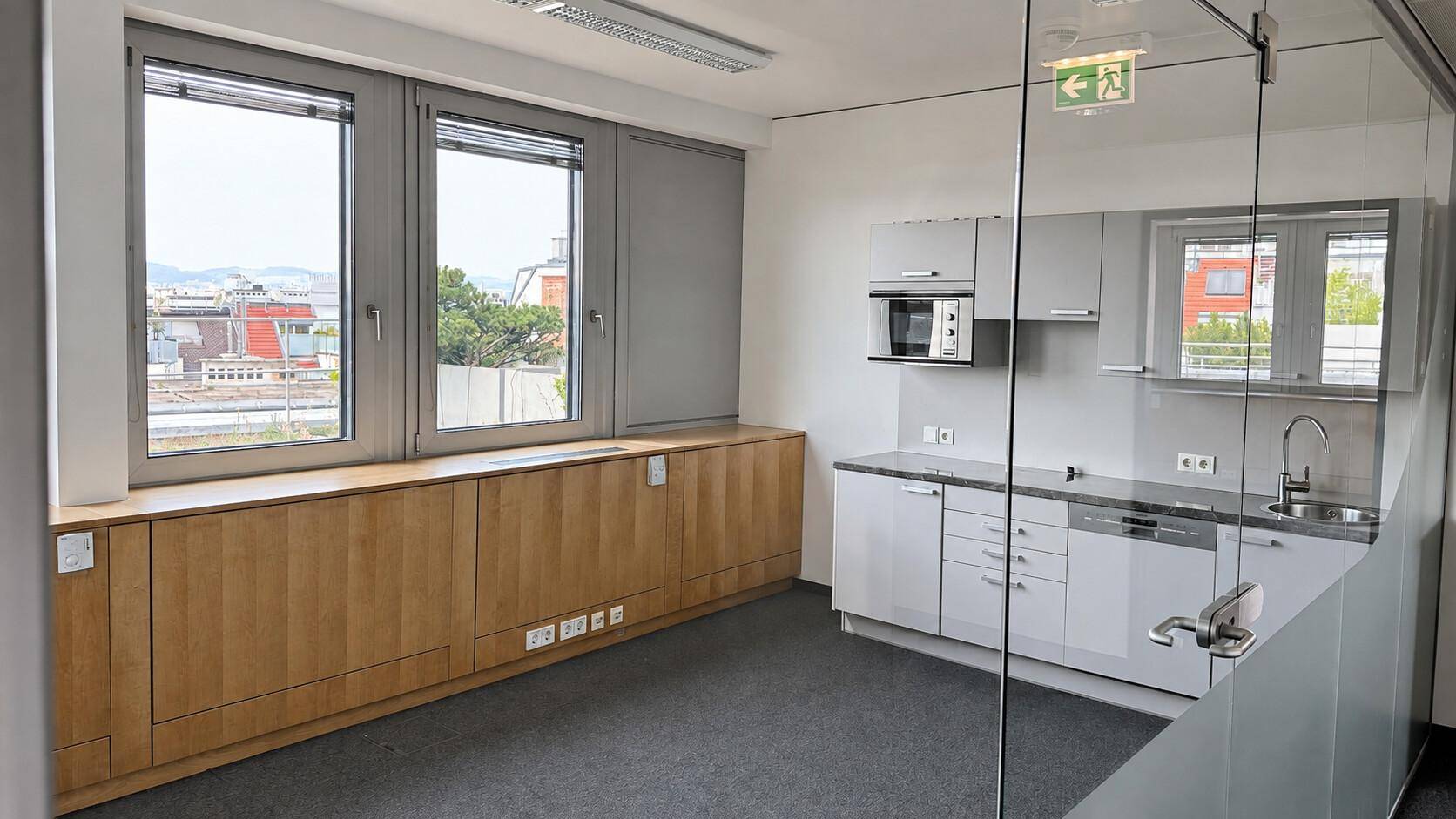 Helles Büro mit integrierter Küchenzeile, Teppichboden und weitem Ausblick durch die Fenster.