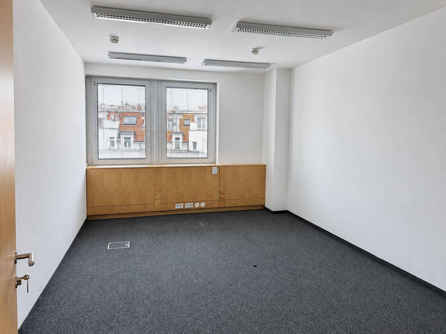Leeres Büro mit Teppichboden, großen Fenstern und integrierten Holzschränken, bereit zur individuellen Gestaltung.