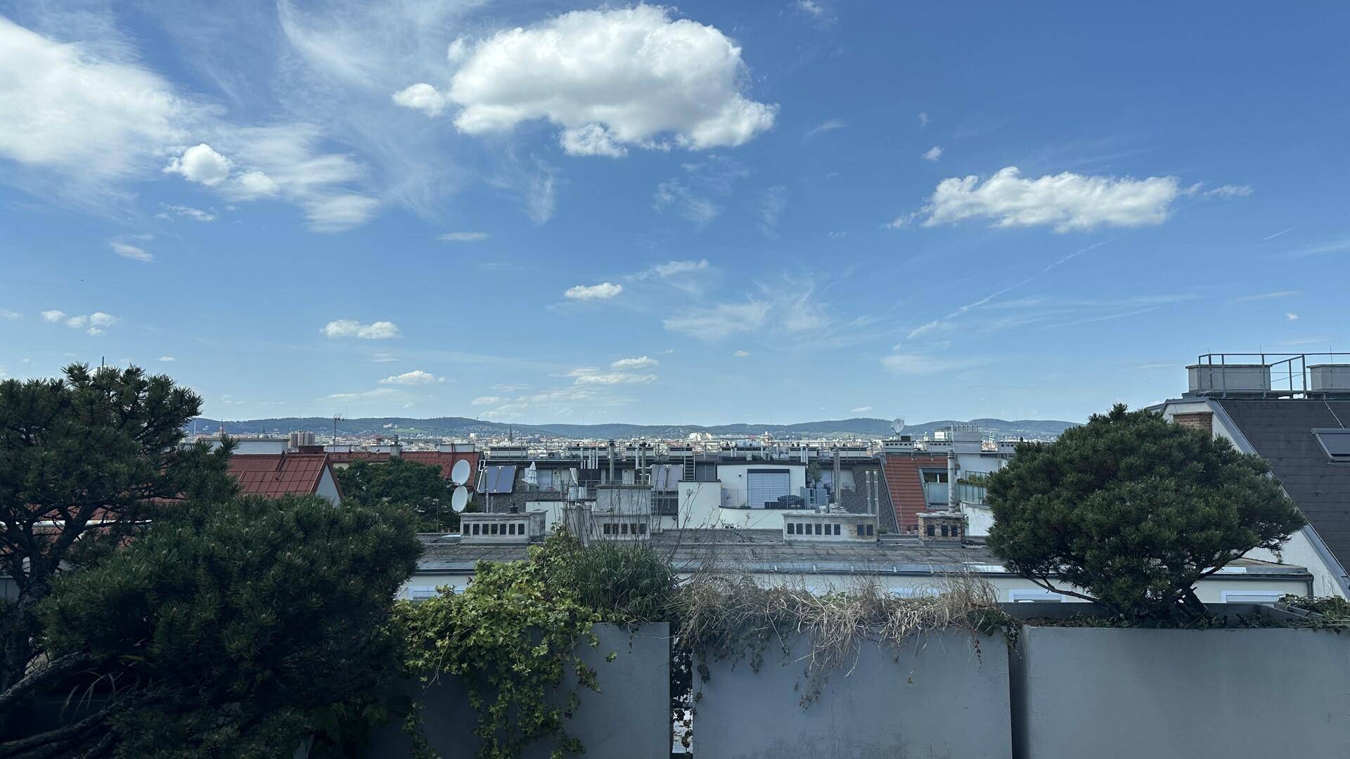 Weitblick über Dächer einer Stadt bis zu den Bergen unter einem klaren blauen Himmel mit Wolken.