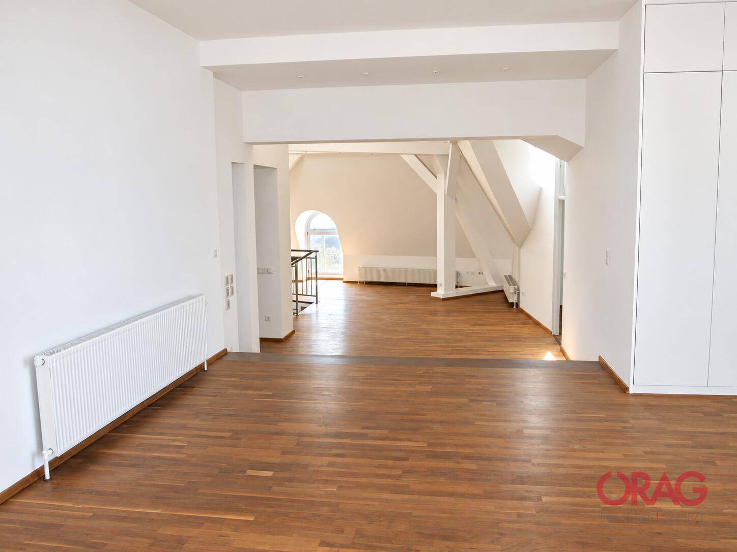 Großer offener Wohnbereich mit Holzboden, weißen Wänden und Blick auf einen Dachgeschossbereich mit Holzbalken.