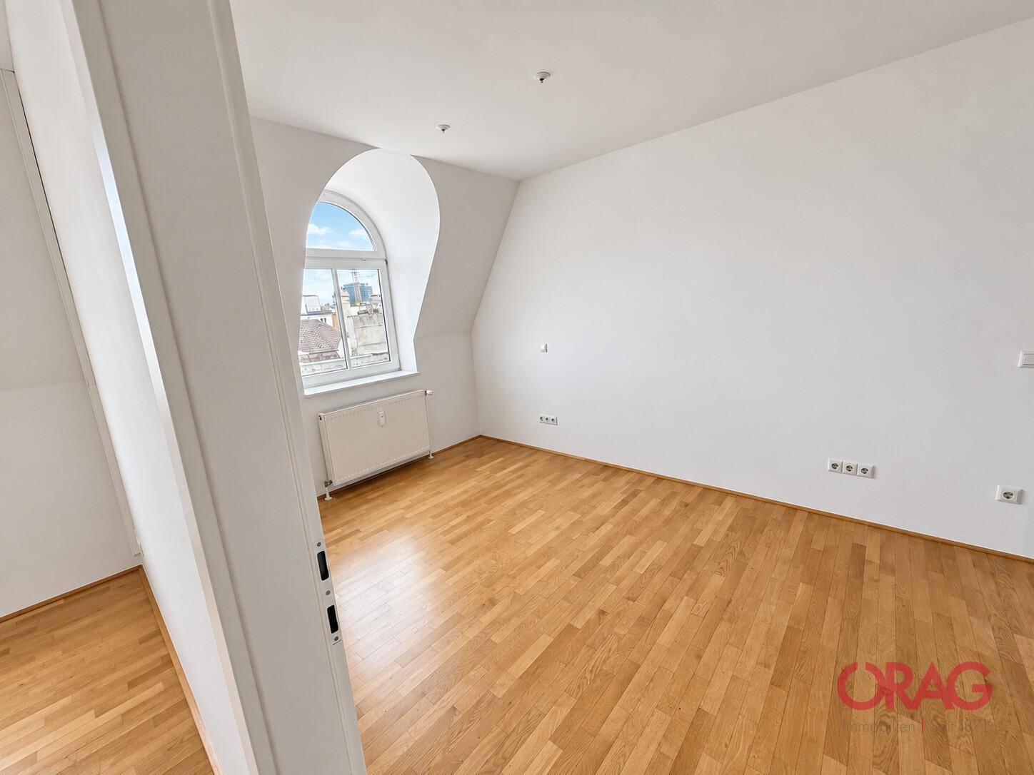 Helles Schlafzimmer mit Holzboden, weißen Wänden und einem charmanten Rundbogenfenster im Dachgeschoss.