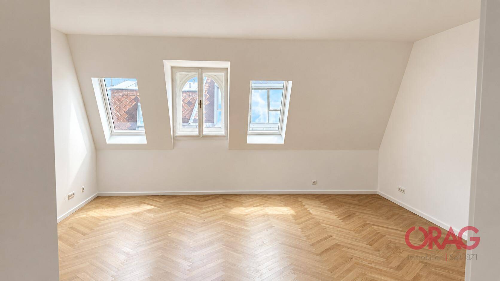 Heller Raum im Dachgeschoss mit schrägen Wänden, Fischgrätparkett und mehreren Dachfenstern.