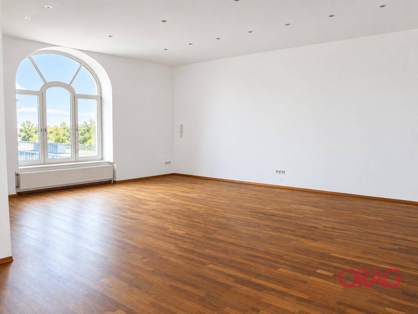 Großer, heller Wohnraum mit Holzboden, weißen Wänden und einem weiten Blick durch das Rundbogenfenster.