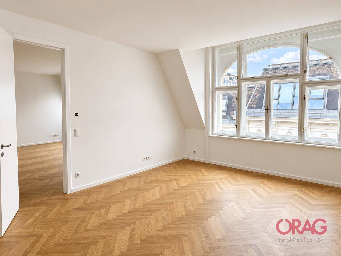 Großer, heller Wohnraum mit Fischgrätparkett, Dachschräge und einer breiten Fensterfront.