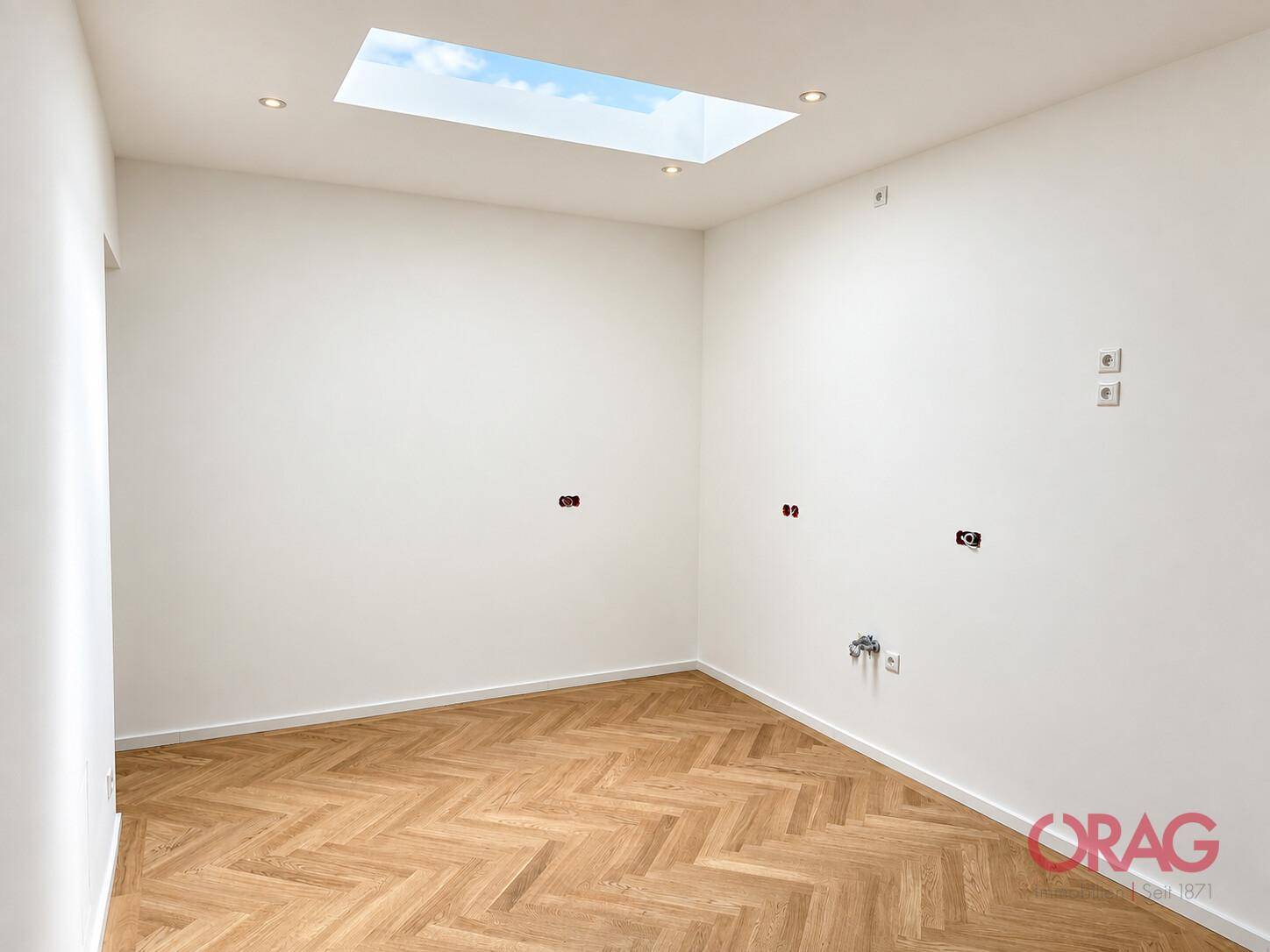 Leerer Raum mit Fischgrätparkett, Deckenstrahlern und einem Oberlicht, vorbereitet für Installationen.