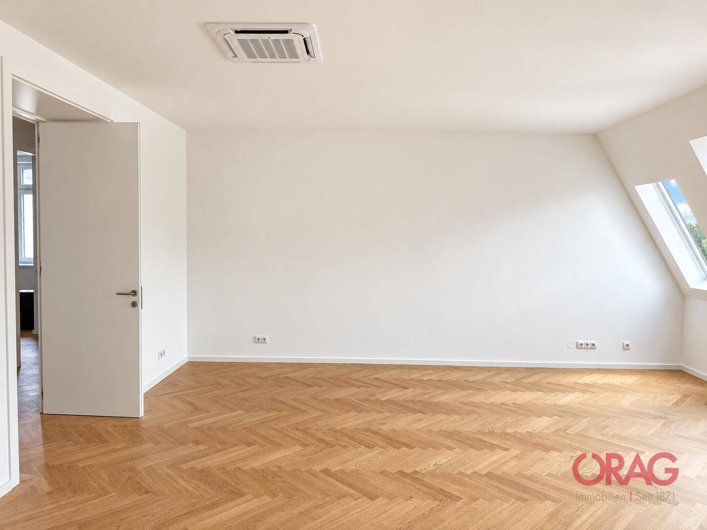 Geräumiger, heller Raum mit Fischgrätparkett, Dachschräge und integrierter Klimaanlage an der Decke.