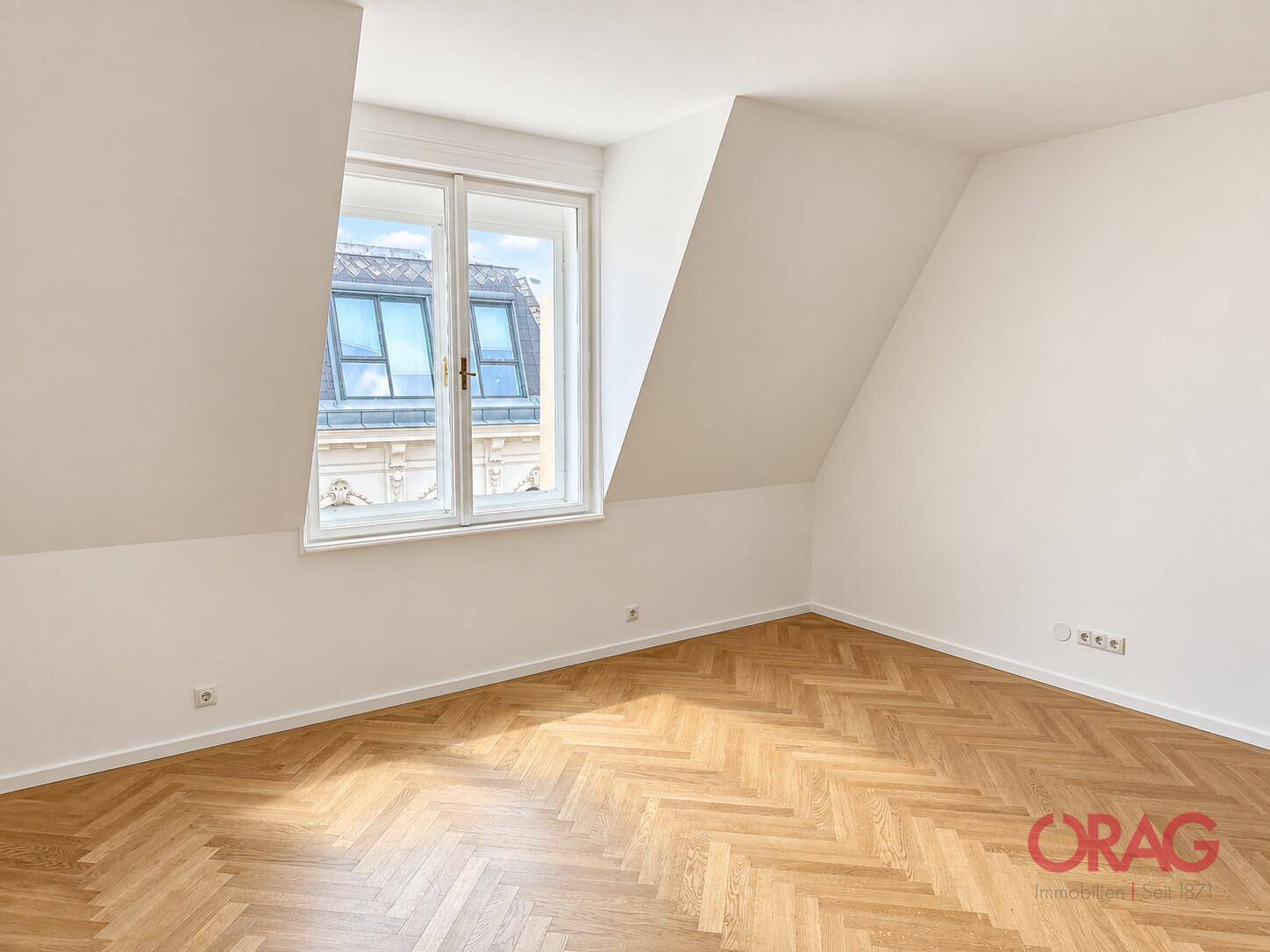Heller Raum im Dachgeschoss mit Fischgrätparkett, schrägen Wänden und einem großen Fenster.