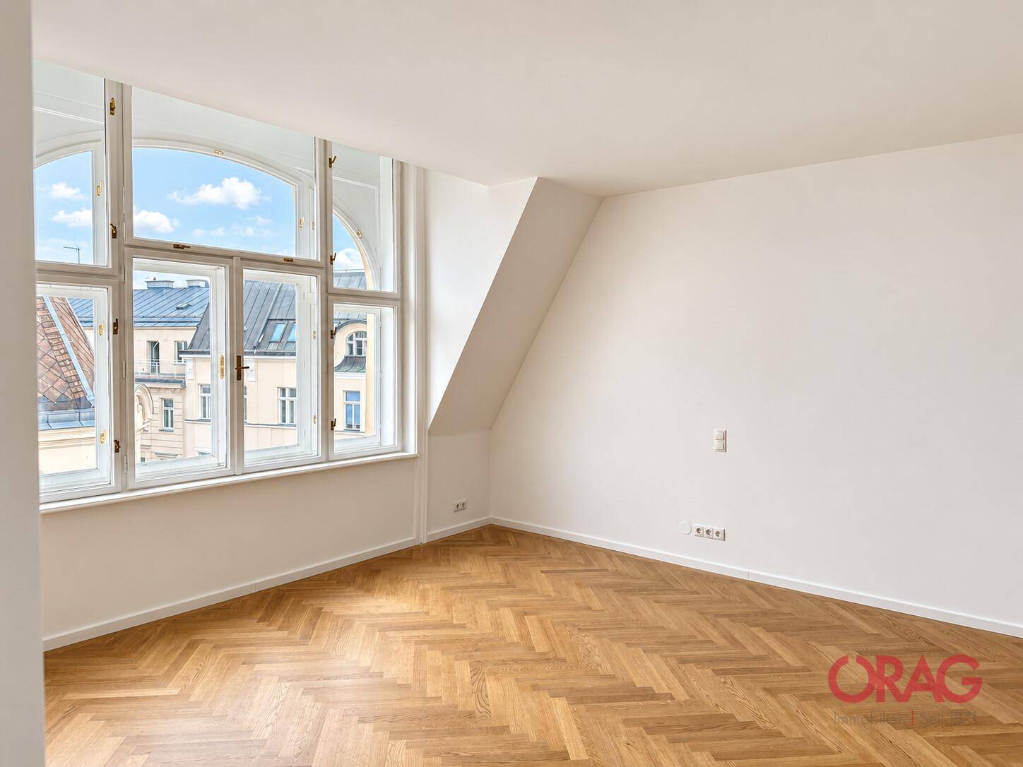 Geräumiger, heller Wohnraum mit Fischgrätparkett, Dachschräge und einer großen Fensterfront.