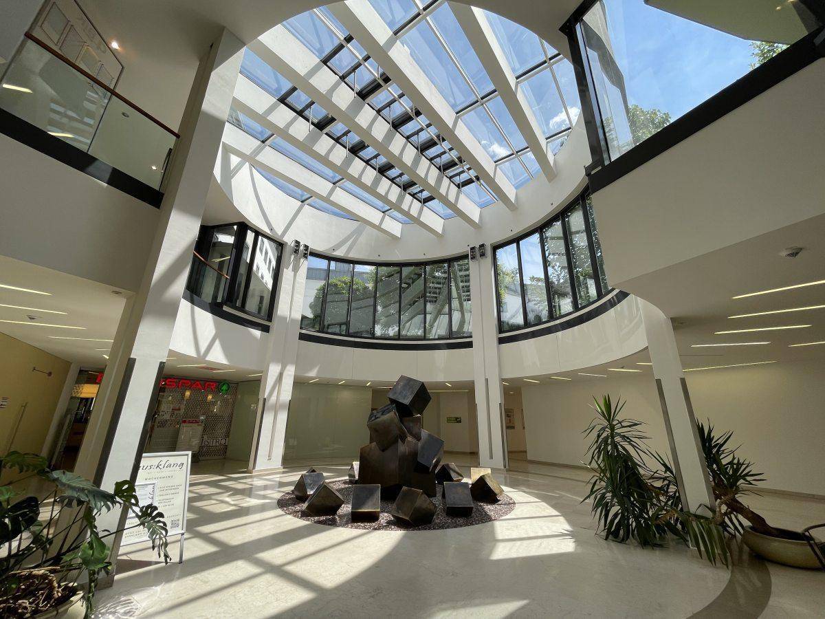 Helles, modernes Atrium mit Glasdach, geschwungenen Wänden und einer zentralen Skulptur, die viel Tageslicht bietet.