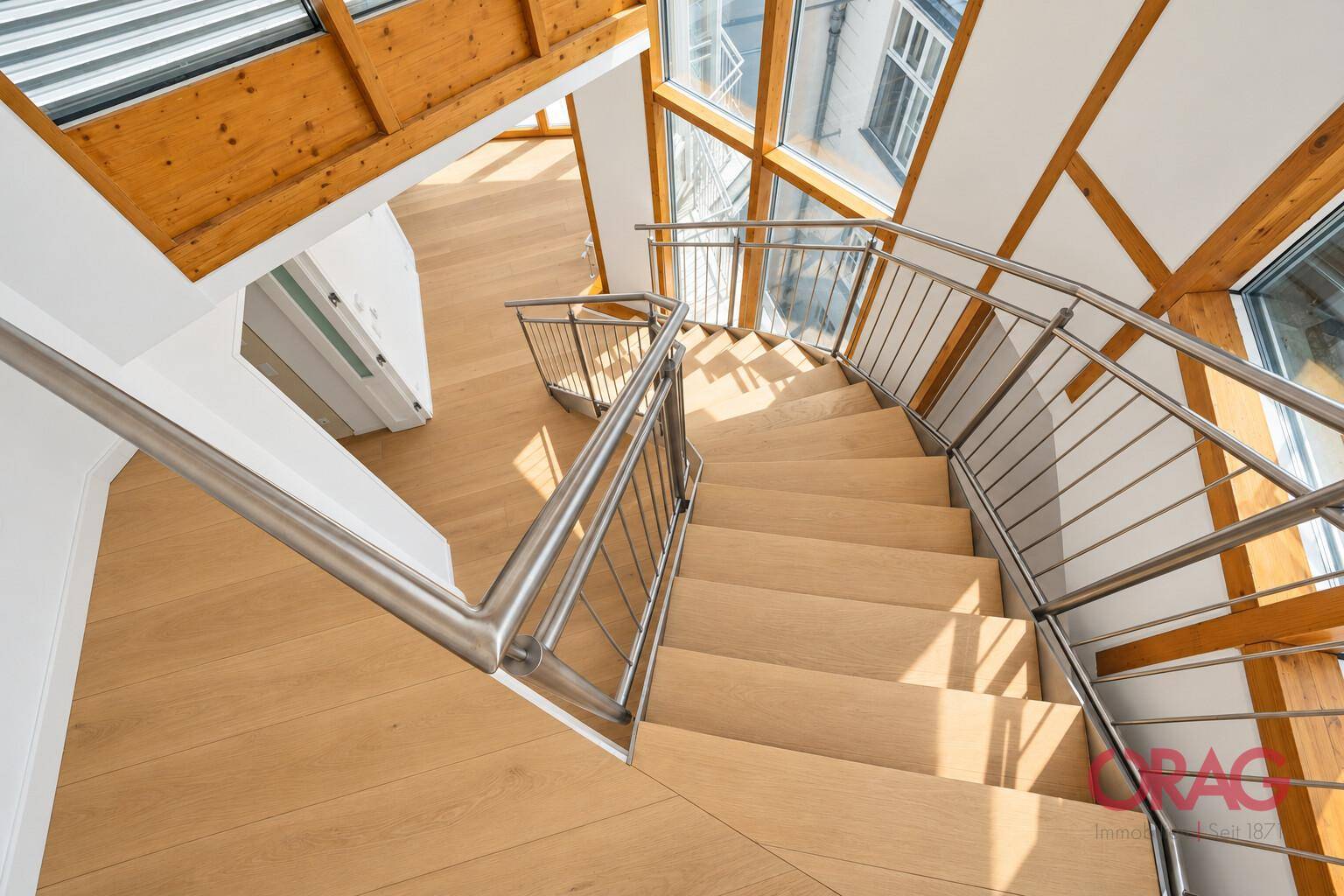 Moderne Holztreppe mit Edelstahlgeländer und großen Fenstern, die viel Licht hereinlassen.