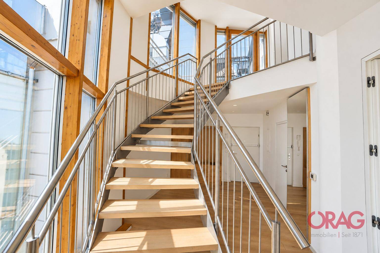 Elegante Holztreppe mit Edelstahlgeländer, umgeben von großen Fenstern, die den Raum hell erleuchten.