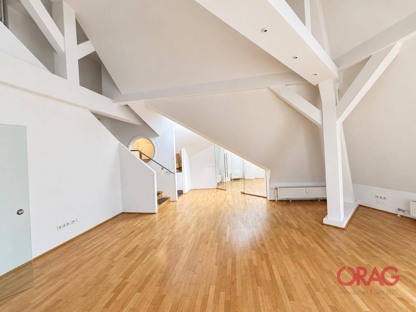 Großer offener Wohnbereich mit sichtbaren weißen Holzbalken, Parkettboden und einer integrierten Treppe.