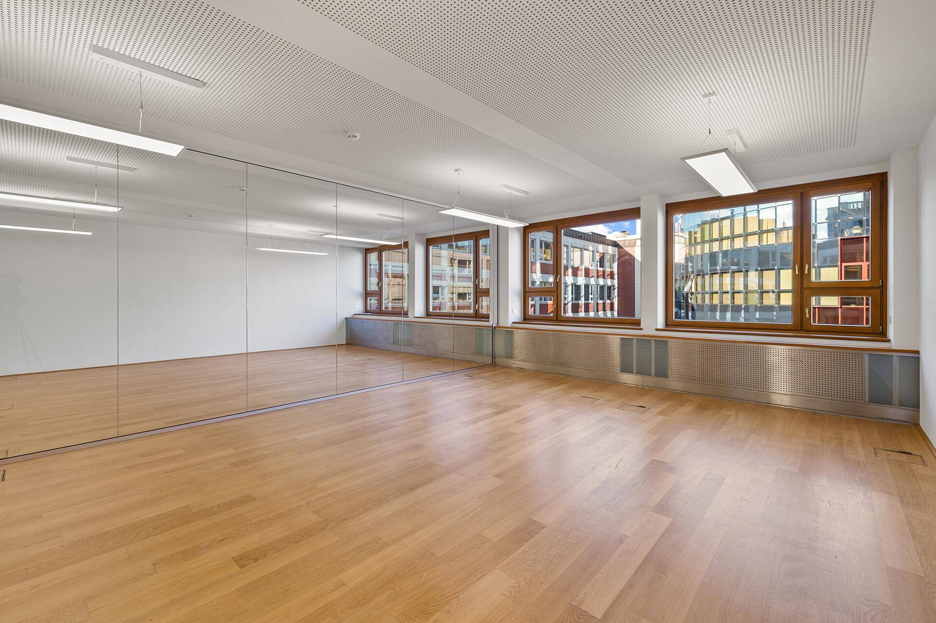 Geräumiger Büroraum mit Holzparkett, großen Fenstern und einer verspiegelten Wand für mehr Weite.