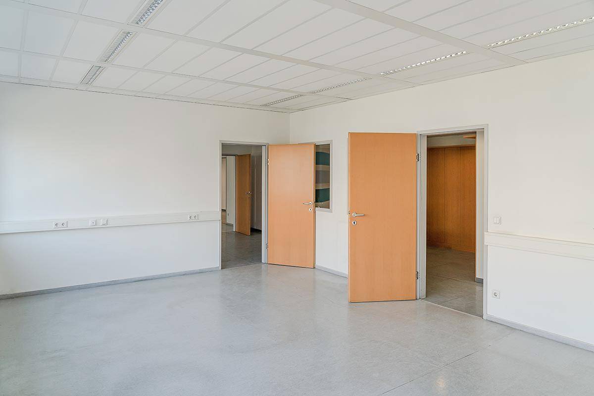 Geräumiger, leerer Raum mit hellem Boden und mehreren Holztüren, die zu angrenzenden Büros führen.
