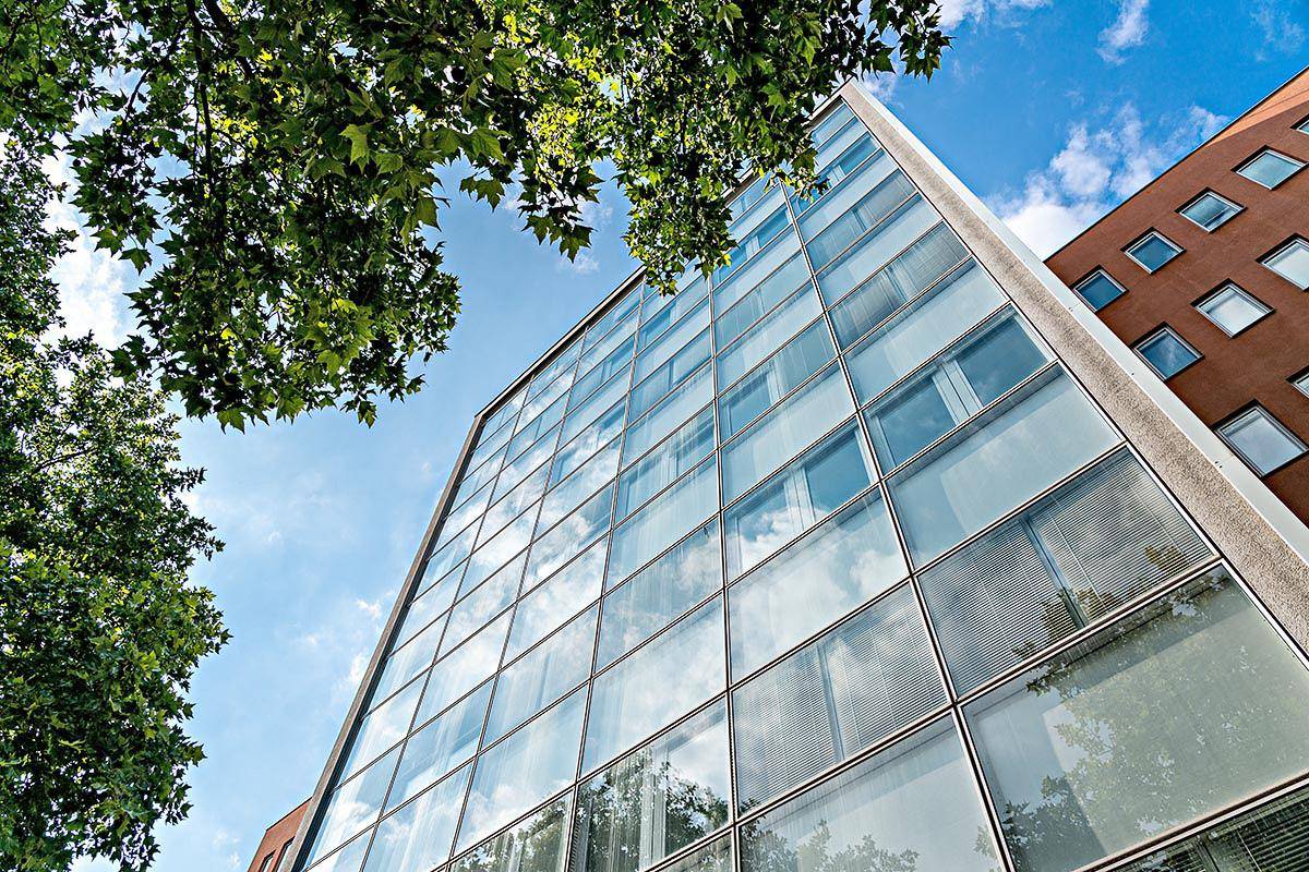 Nahaufnahme einer modernen Glasfassade, die den blauen Himmel und Bäume widerspiegelt, mit einem angrenzenden Gebäude.