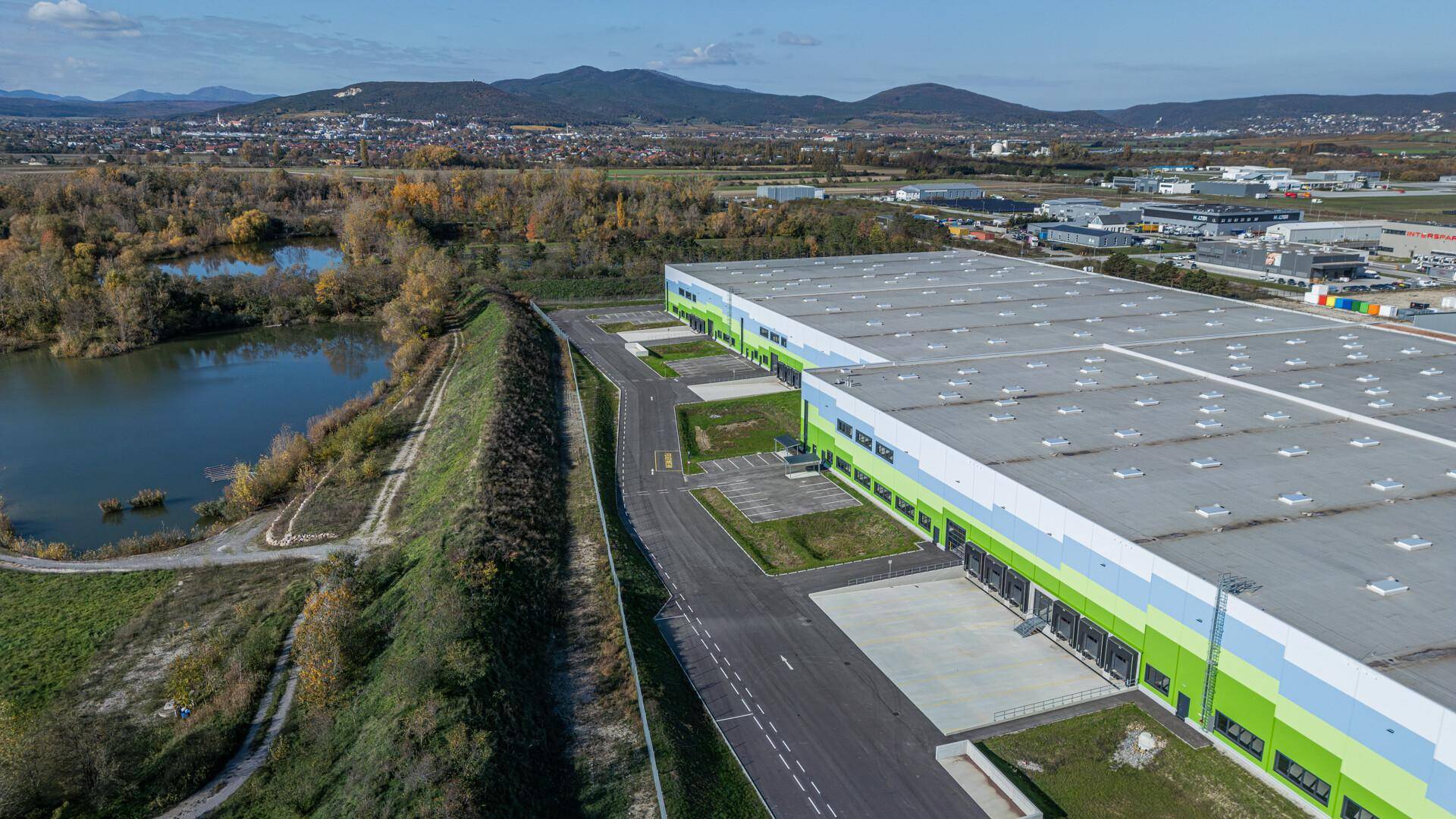 Große Industriehalle neben einem See, umgeben von grüner Landschaft und Bergen unter blauem Himmel.