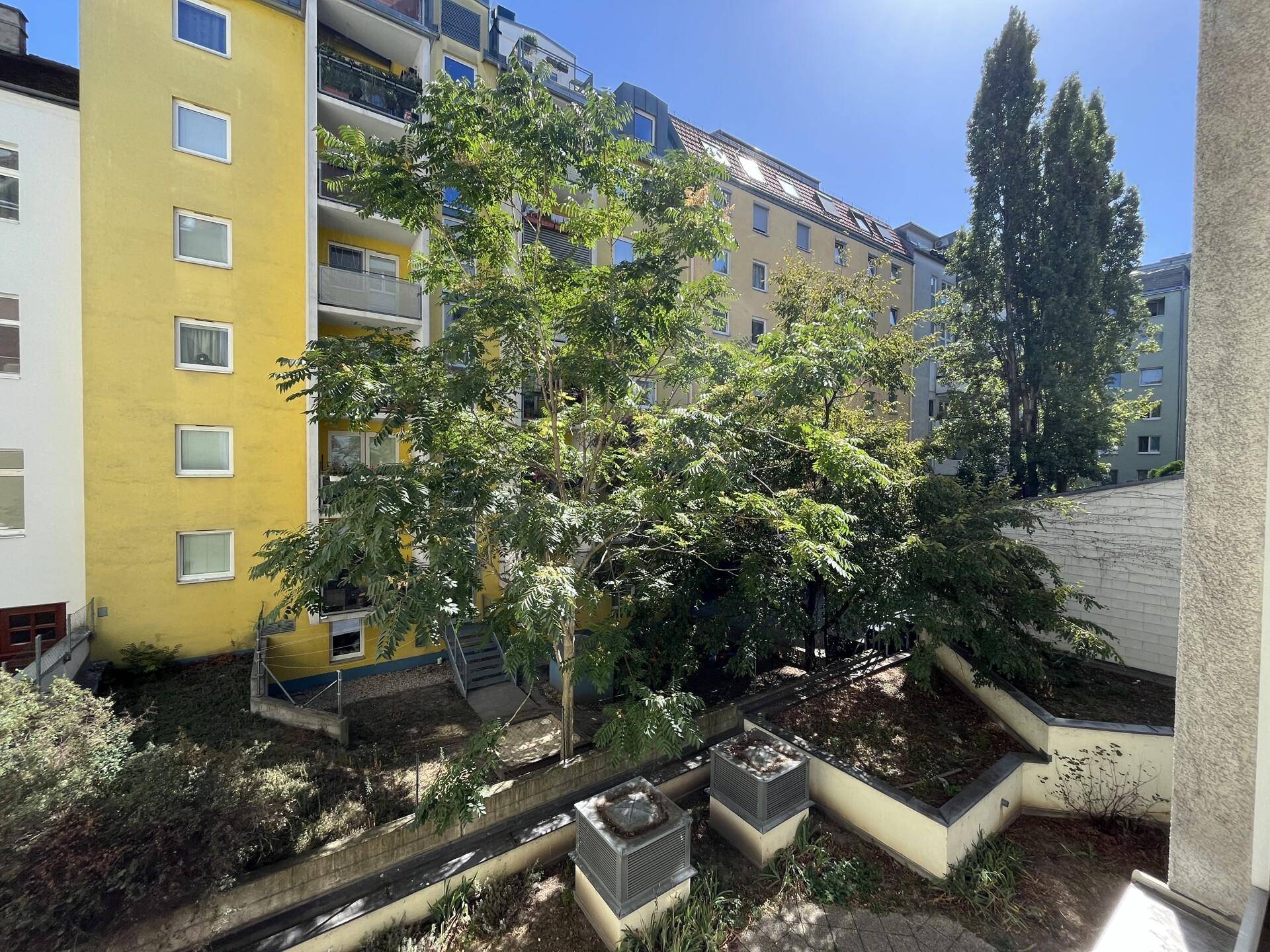 Blick von einem Balkon in einen grünen Innenhof mit Bäumen und angrenzenden Gebäudefassaden.