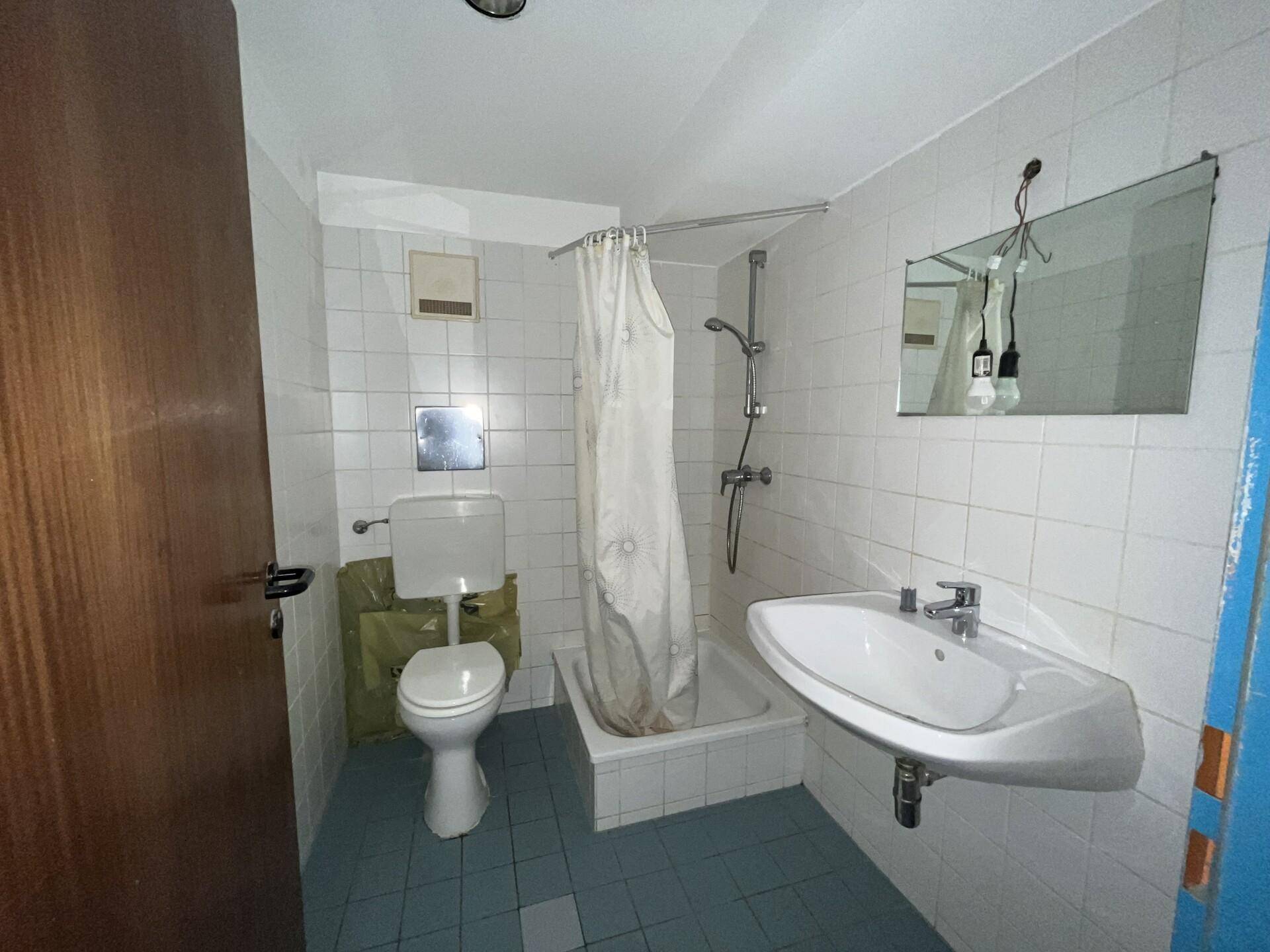 Badezimmer mit weißen Wandfliesen, blauen Bodenfliesen, Toilette, Waschbecken und Duschbereich mit Vorhang.