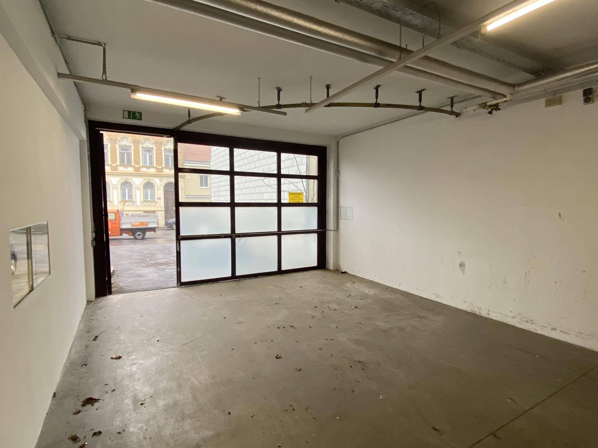 Leerer Garagenraum mit großem Glasrolltor zur Straße und sichtbaren Deckeninstallationen.