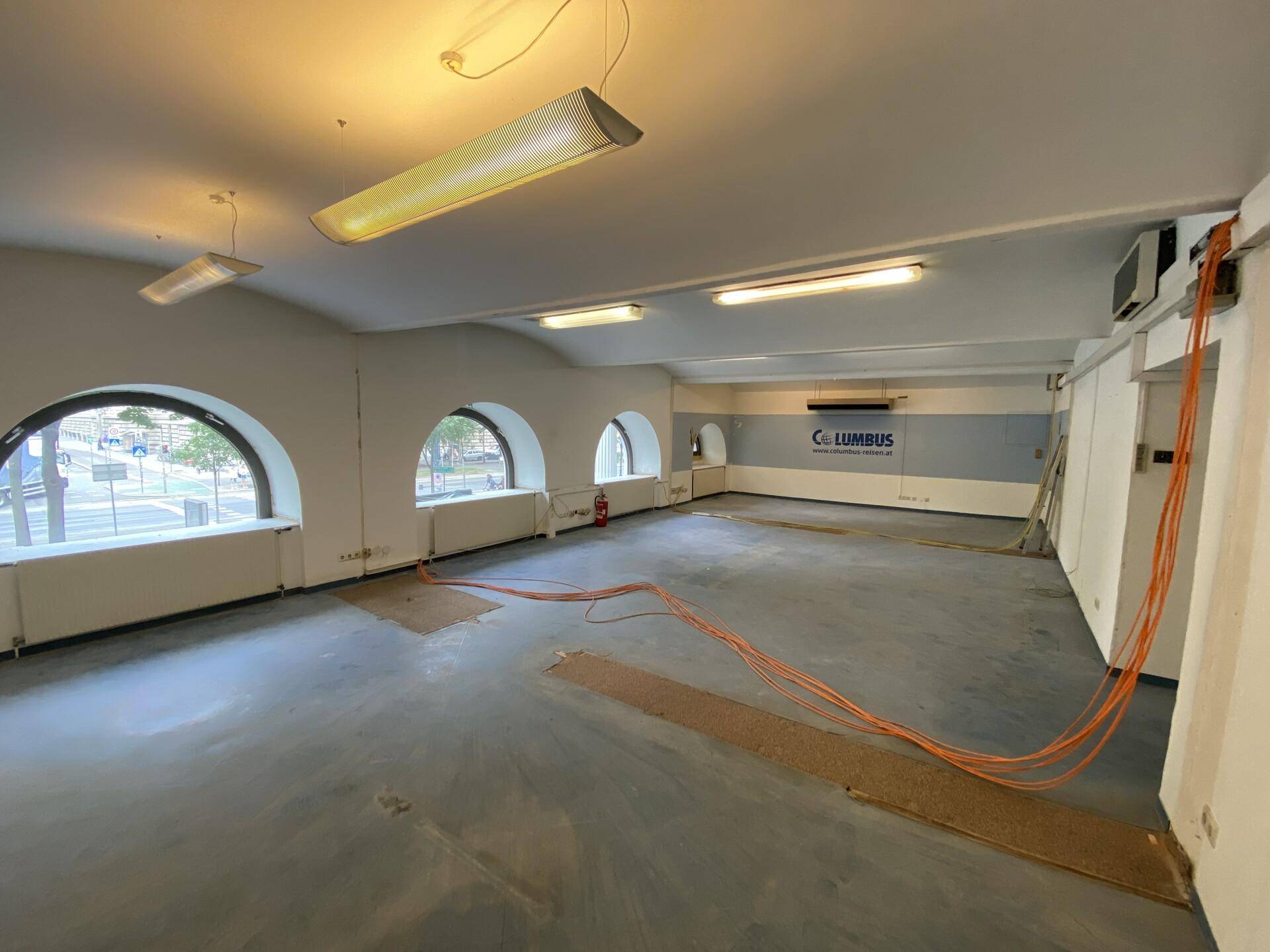 Geräumiger Büroraum mit großen Rundbogenfenstern, Betonboden und sichtbaren Installationen.