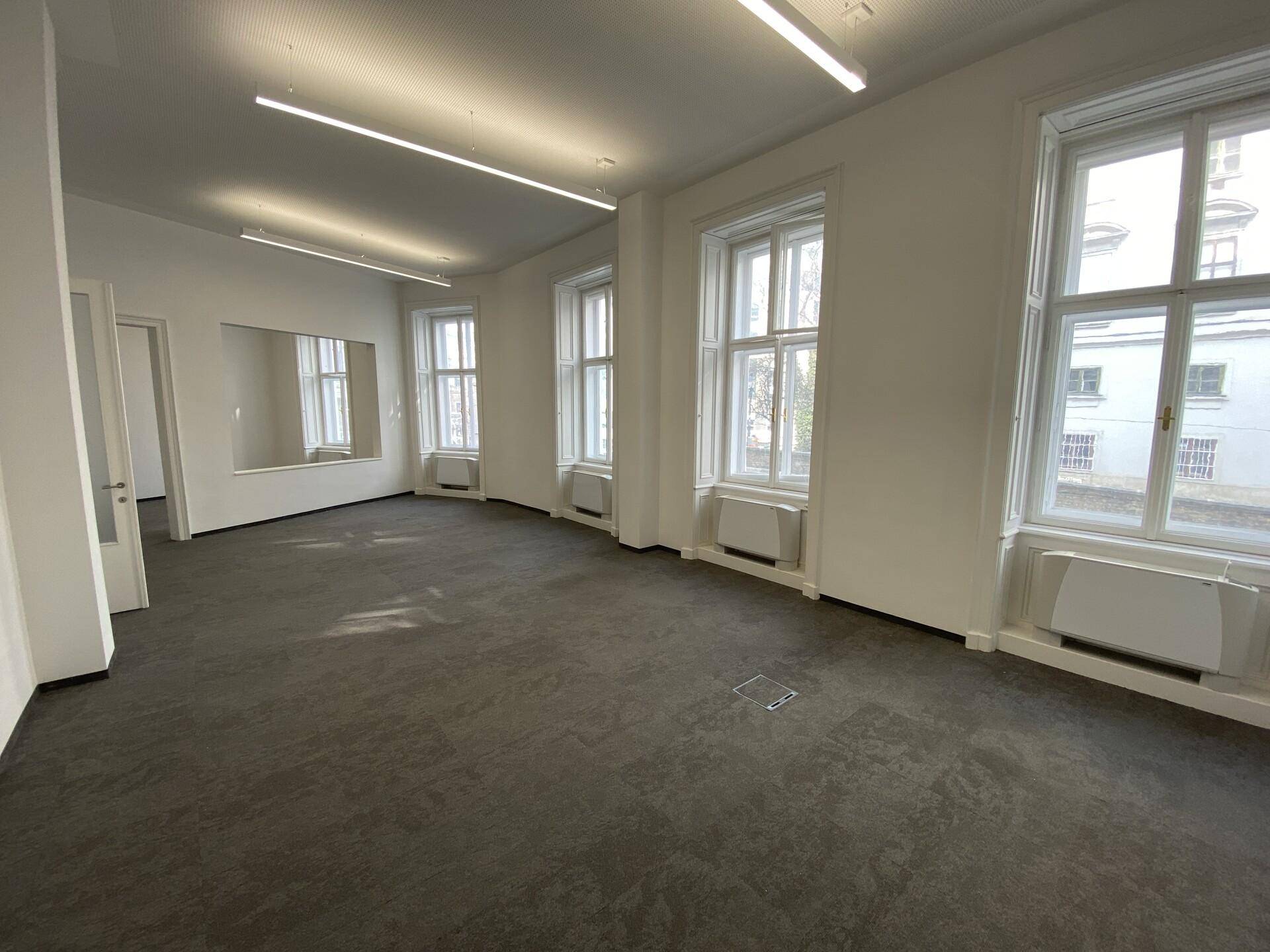 Heller Büroraum mit Teppichboden, großen Fenstern und integrierten Heizkörpern unter den Fenstern.