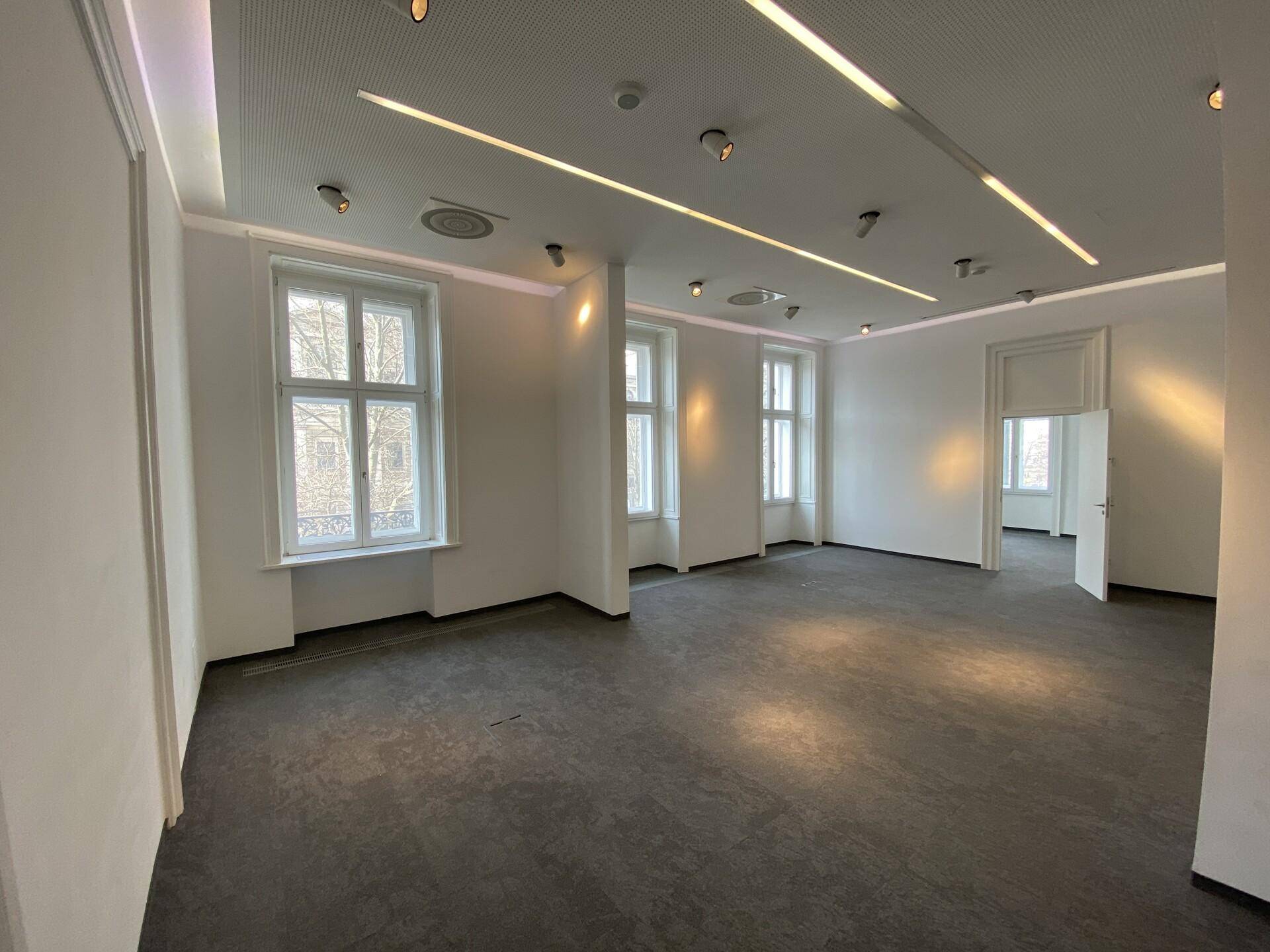 Geräumiger Büroraum mit dunklem Boden, großen Fenstern und moderner Deckenbeleuchtung.