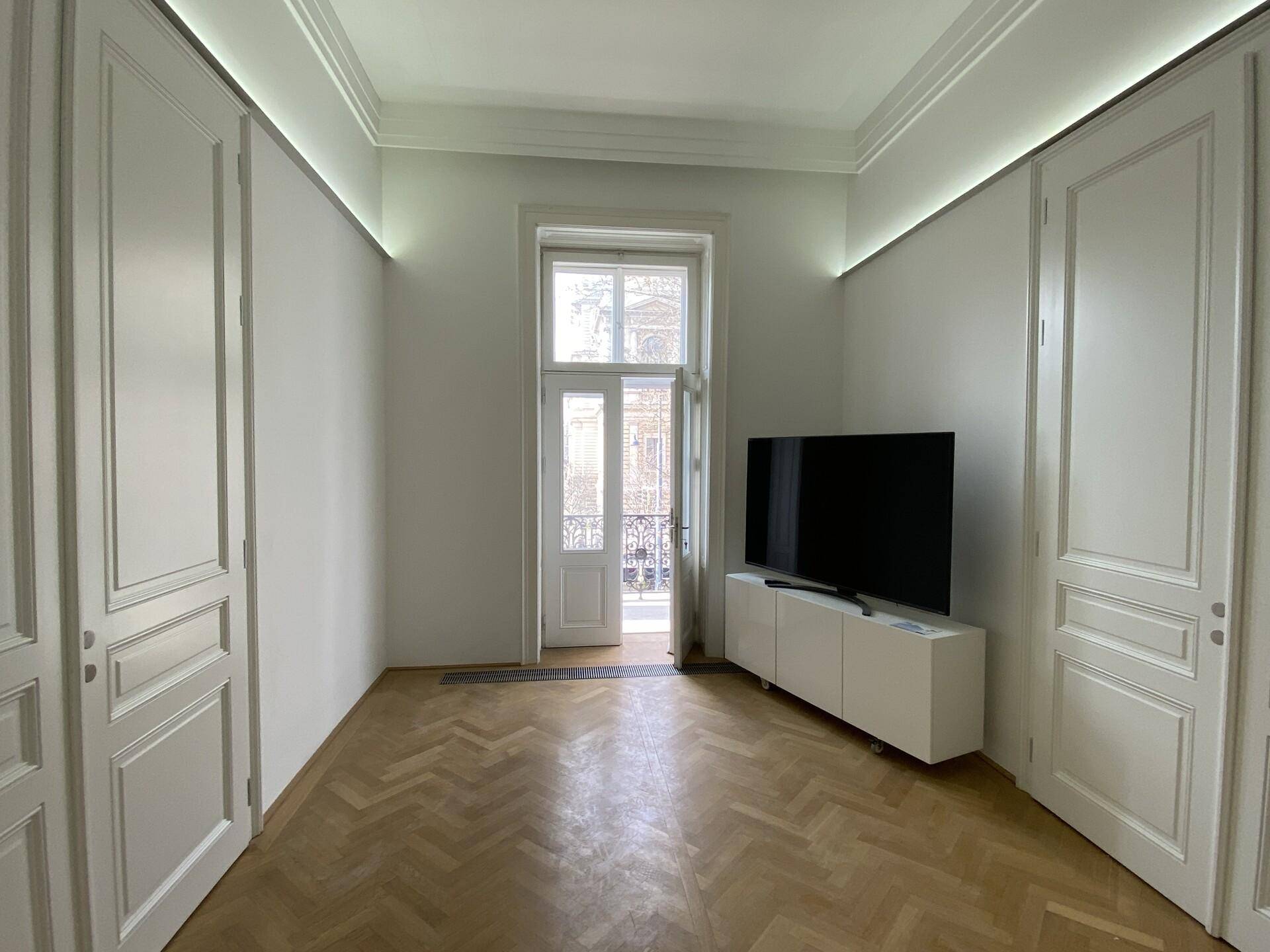 Heller Raum mit Fischgrätparkett, hohen Türen und einem französischen Balkon mit Blick nach draußen.