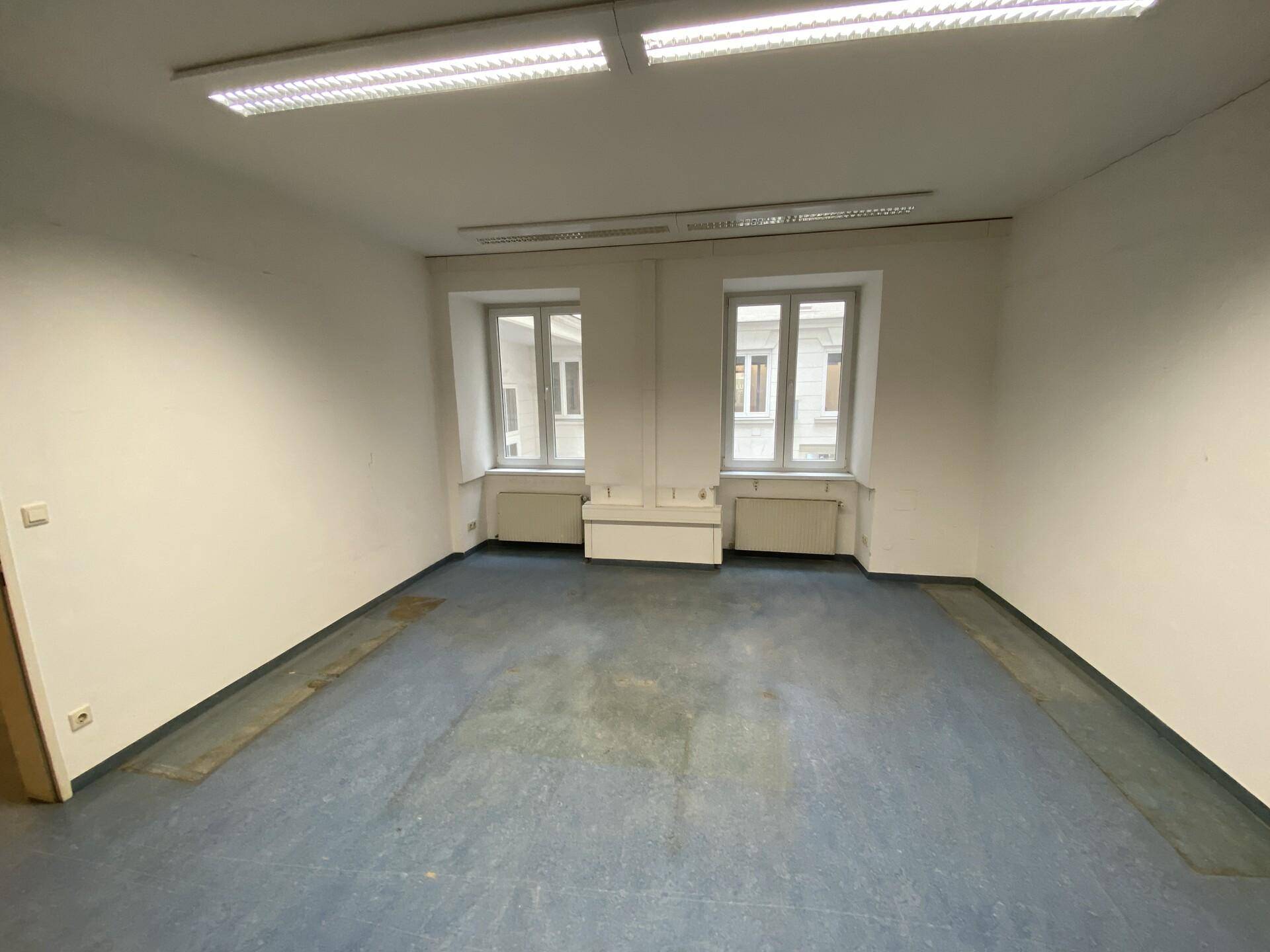 Leerer Büroraum mit zwei Fenstern, Heizkörpern und einfacher Beleuchtung.